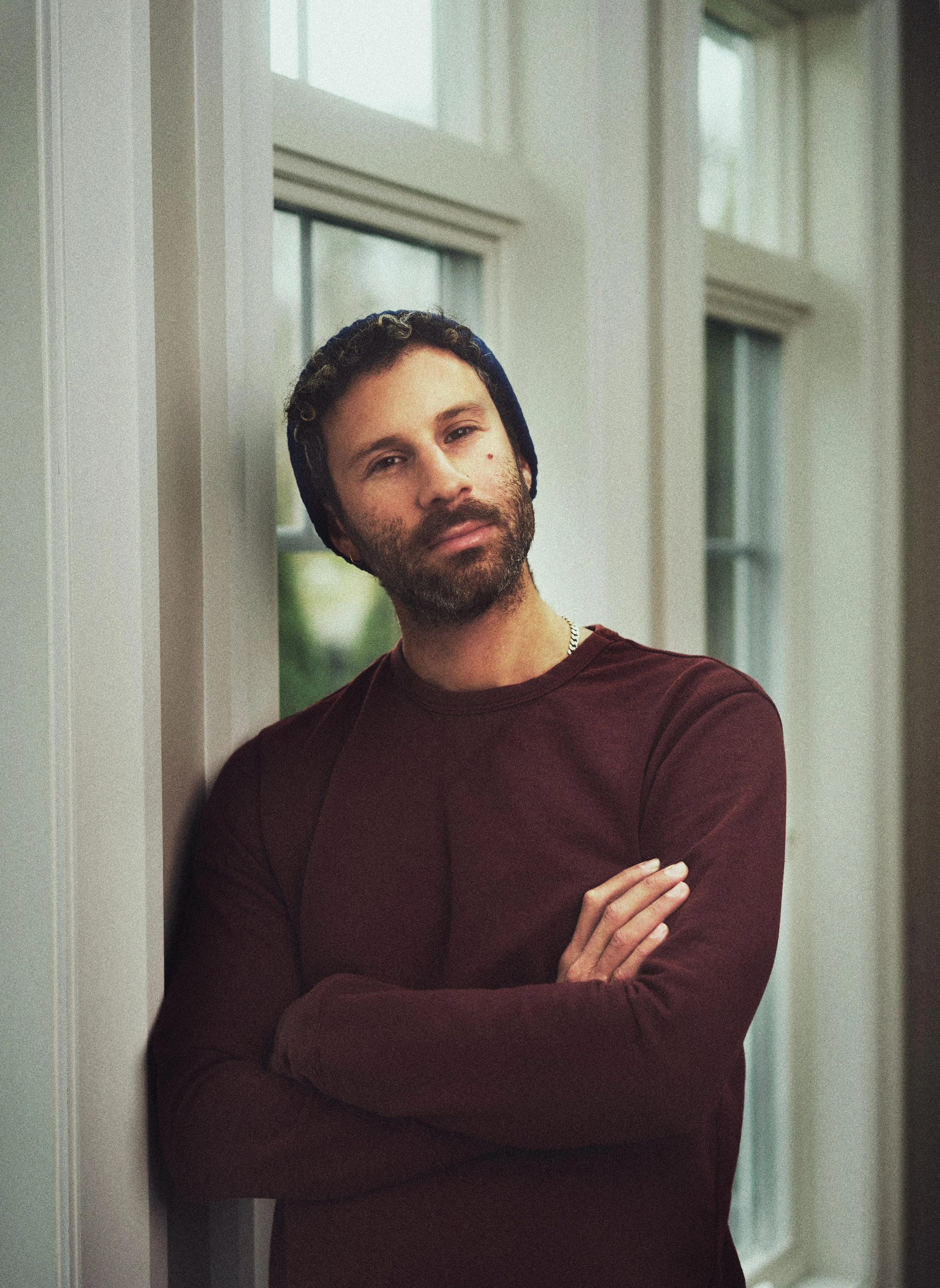 A man with a beard, wearing a dark blue beanie and maroon long-sleeve shirt, leaning against a wall near large windows with white frames, arms crossed, looking at the camera.