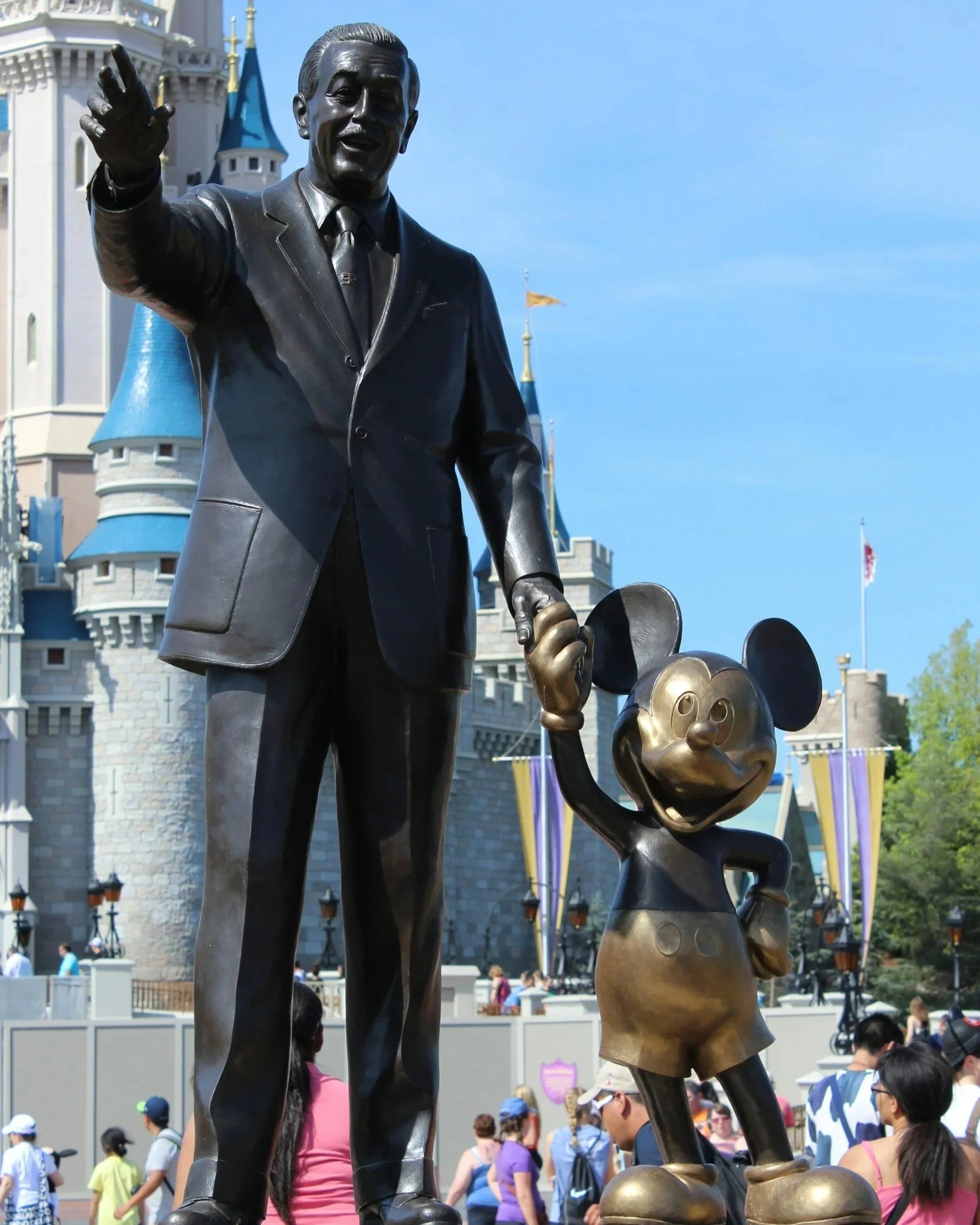 Statue of Walt Disney holding Mickey Mouse's hand in front of a Disney castle at a Disney theme park.