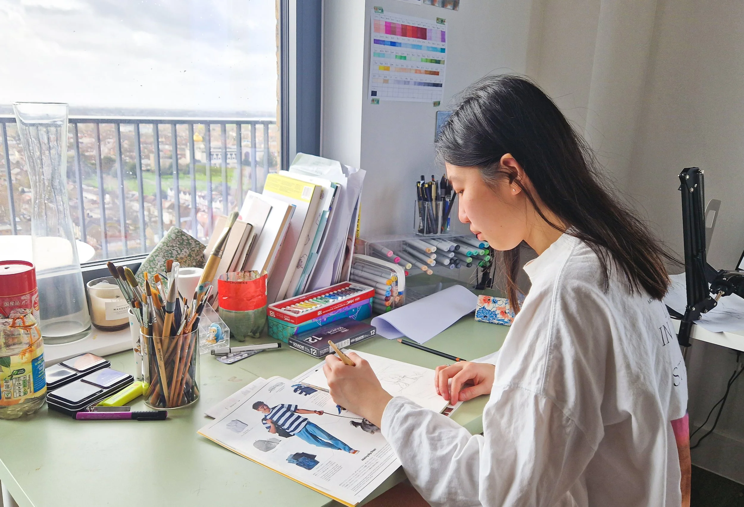 A woman with dark hair is seated at a cluttered desk, drawing or writing in a magazine or catalog with fashion images. The desk is filled with art supplies, including paint brushes in containers, markers, and pens. There is a window with a view of a cityscape and an overcast sky in the background. Behind her, a colorful chart and some papers are taped to the wall.