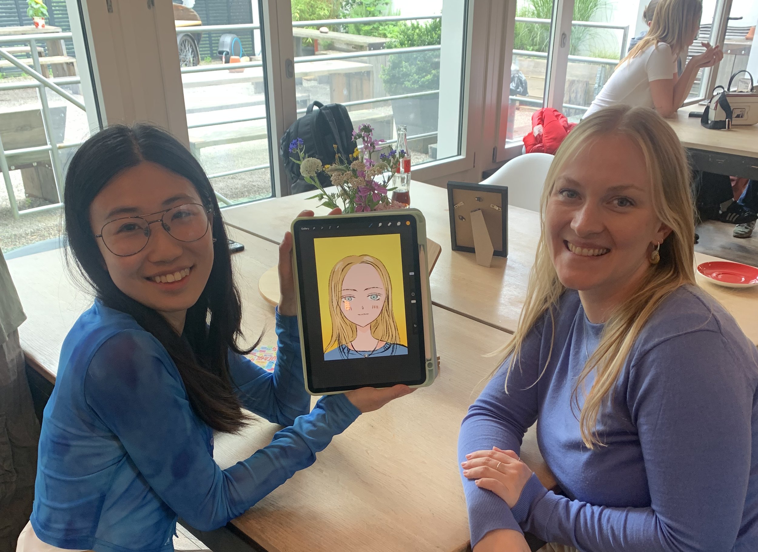 Two women sitting at a table in a bright indoor space. The woman on the left, with black hair and glasses, is holding a tablet showing a cartoon portrait of a woman with blonde hair and blue eyes. The woman on the right, with blonde hair, is smiling 