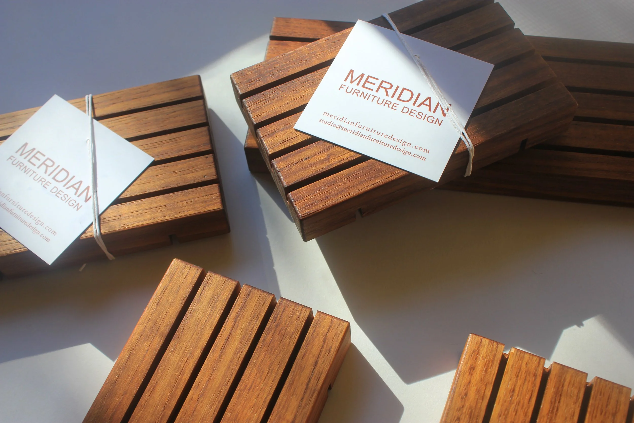 teak wooden soap trays in assorted sizes shown unwrapped alongside some in packaging with Meridian Furniture Design logo in red text on white label.