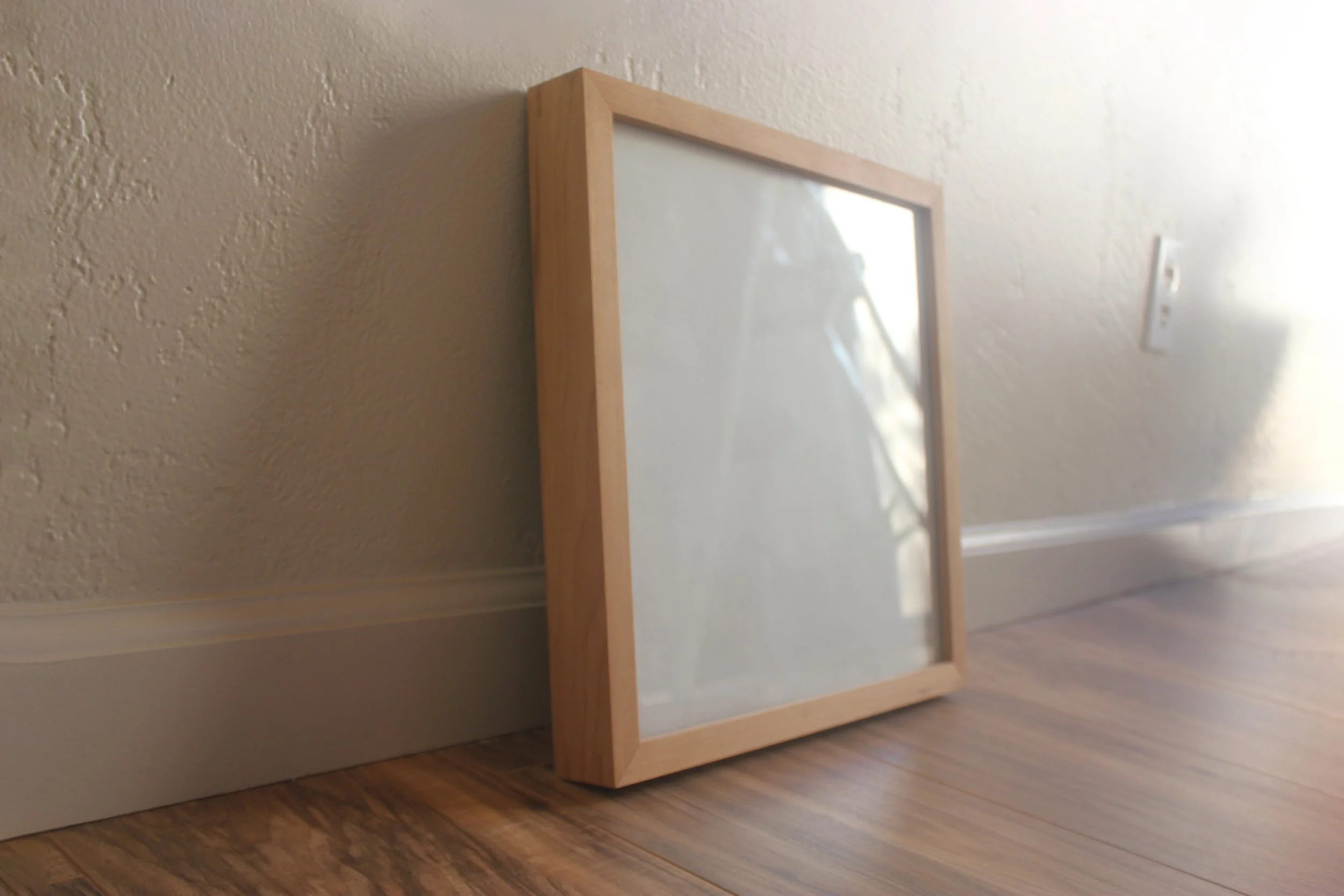 A 12" x 12" x 1.75" gallery frame made of solid cherry wood with clear glass. Shown in a home setting against a white wall.