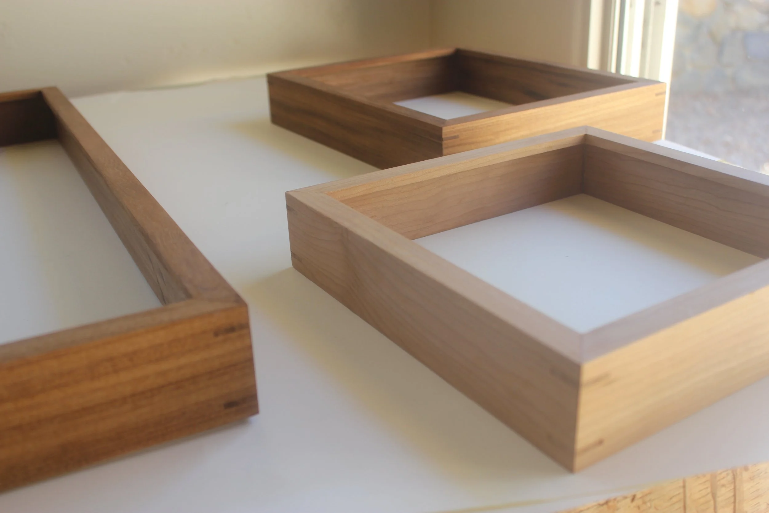 Three gallery frames shown on a work desk in a home setting. Frames shown are made of teak and cherry.