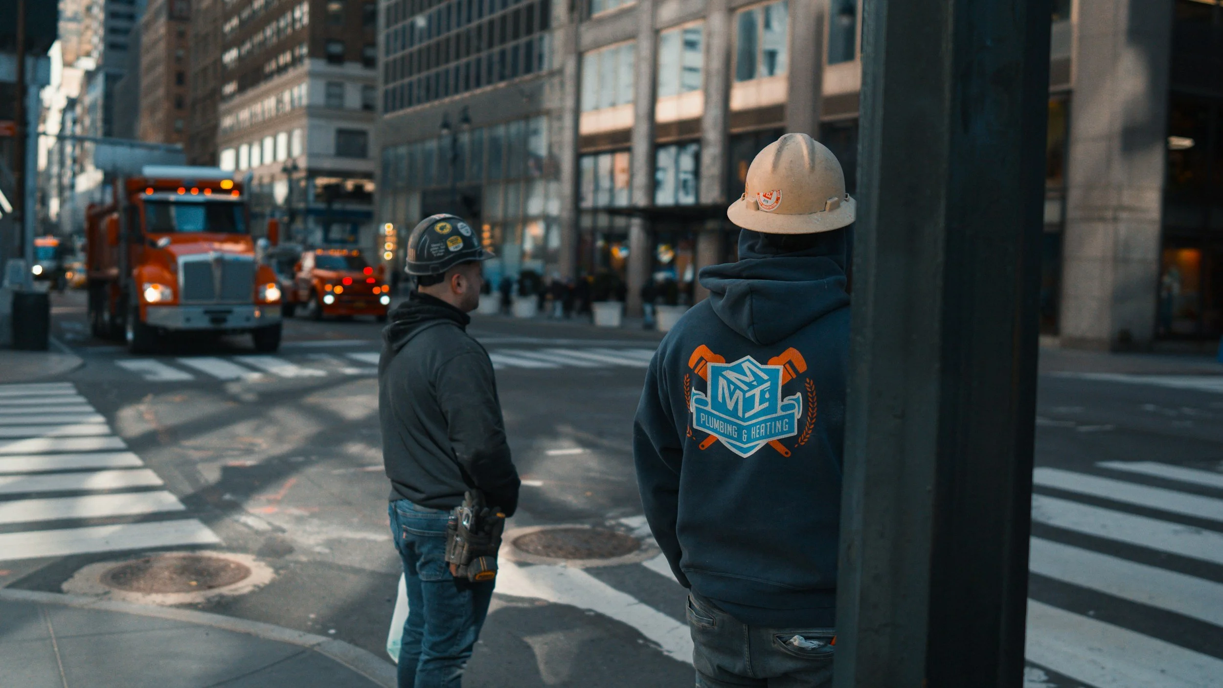 Two construction workers with hard hats standing at a city crosswalk, with trucks and tall buildings in the background.