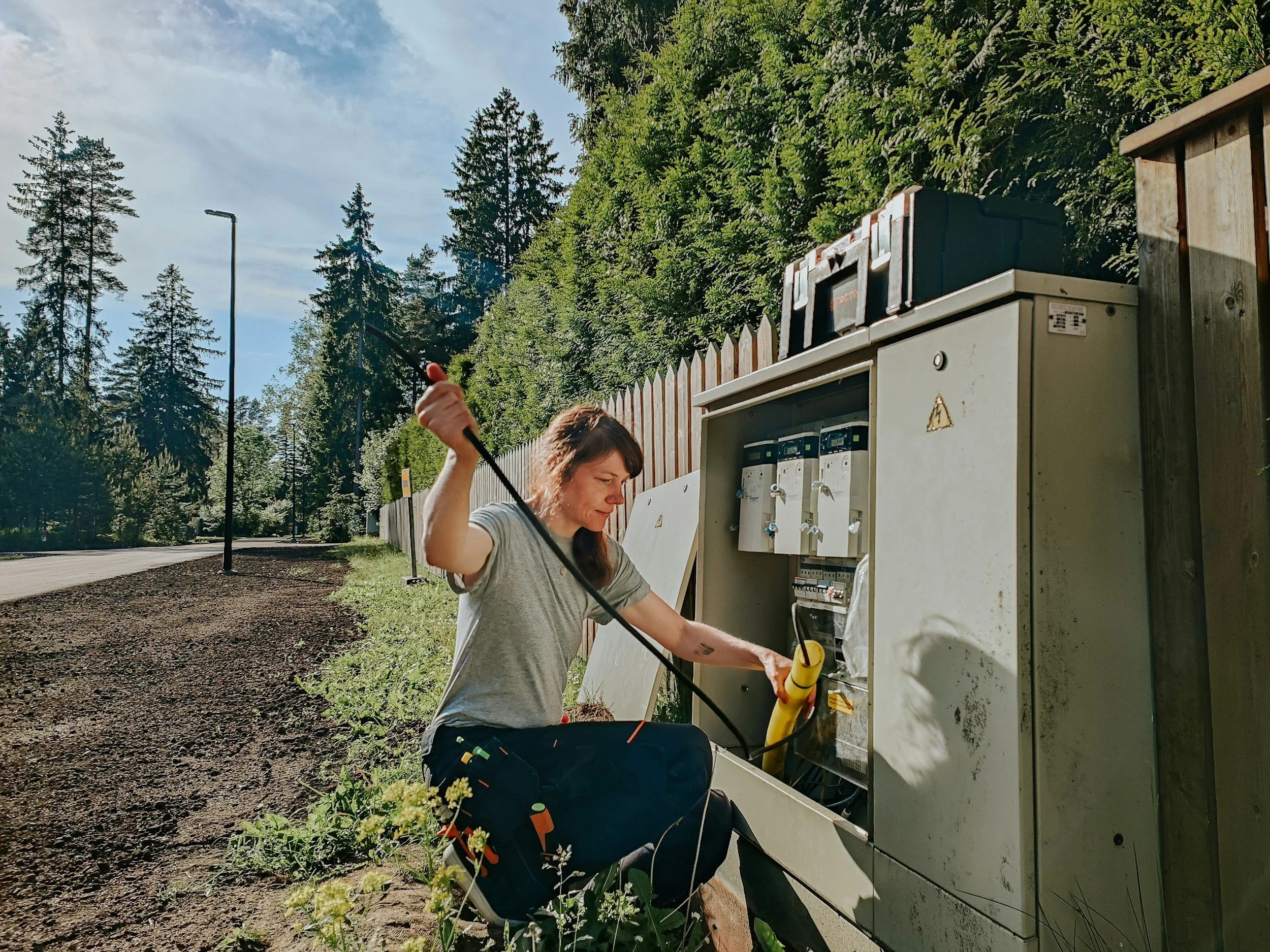 A woman working on a utility box along a forested roadside, holding a tool and wearing casual clothing.