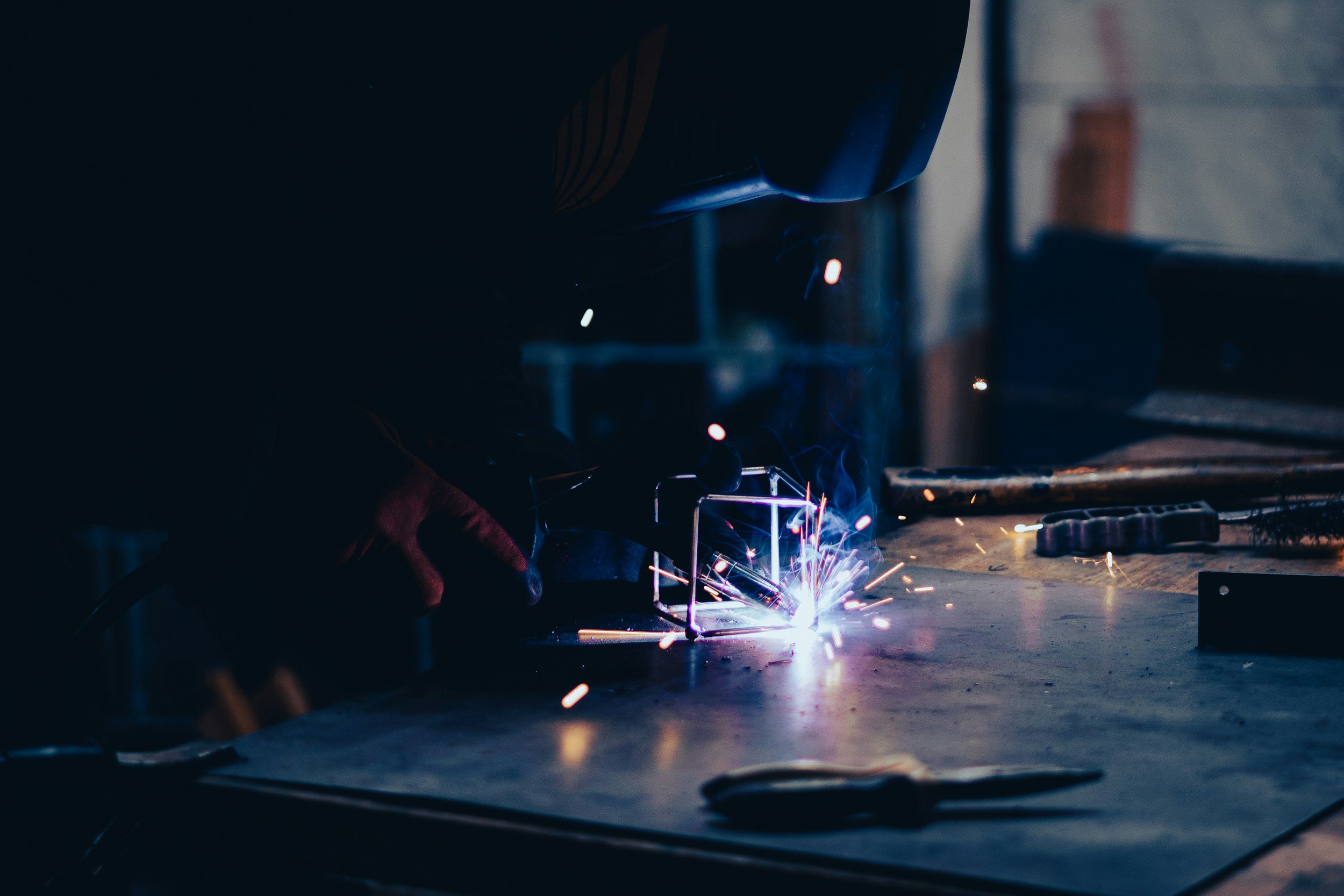 A person welding metal pieces on a workbench in a workshop with sparks flying.