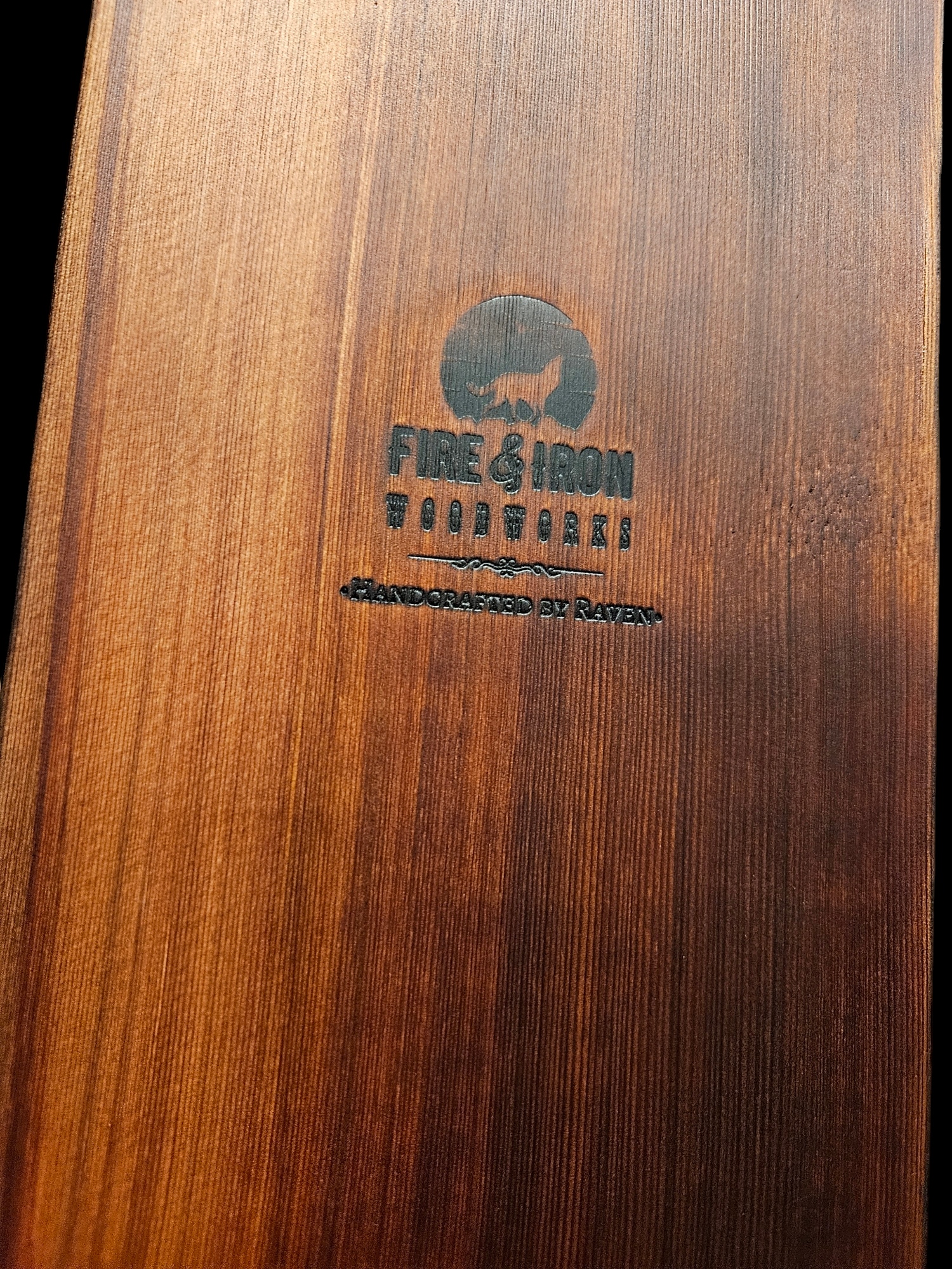 Close-up of a wooden surface with a logo featuring a lion silhouette and the text 'Fire & Iron' and 'Toudoukei Handcrafted by Raven' engraved or printed on it.