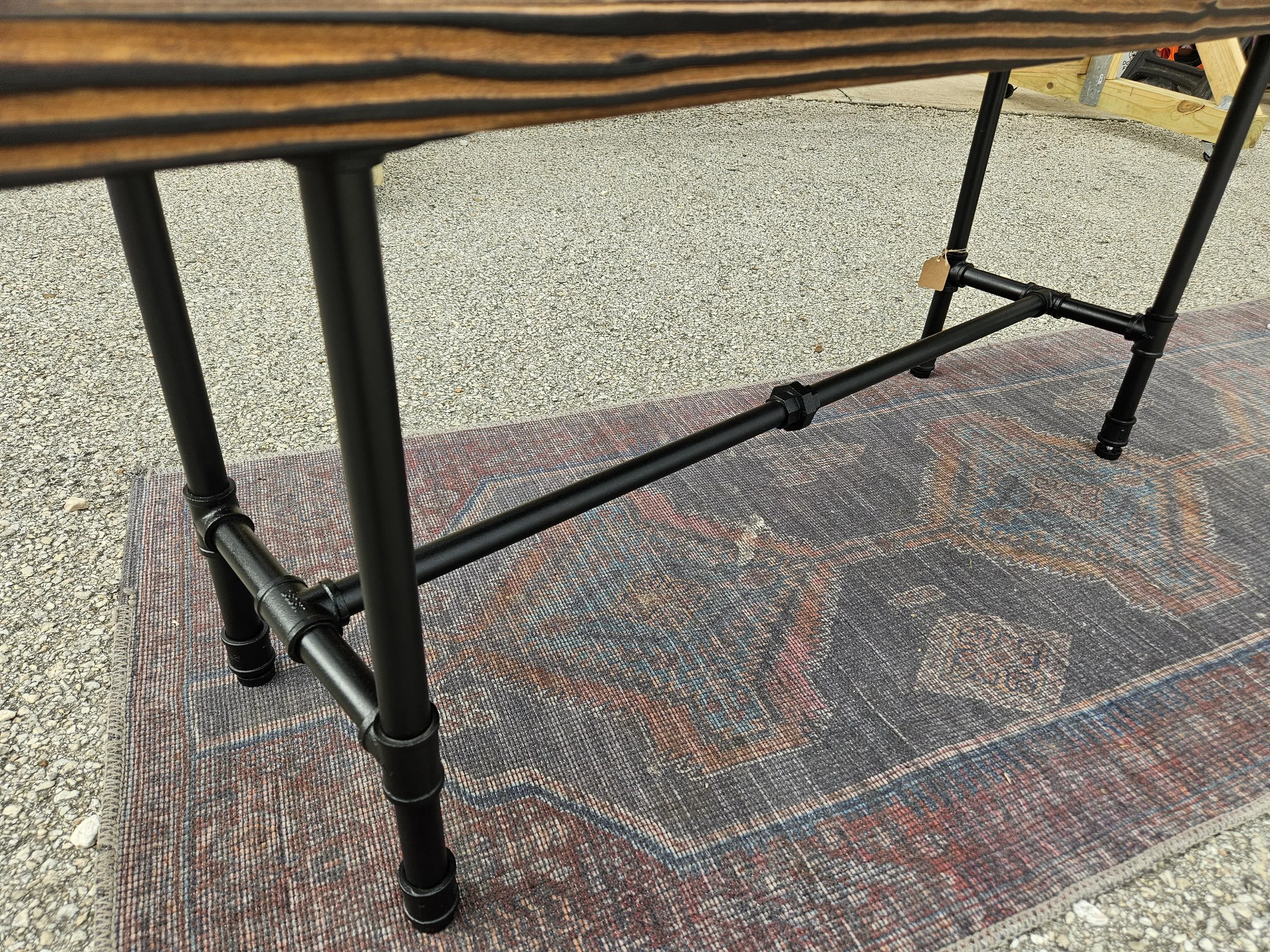 A close-up of a black metal table frame with wooden top and a tag hanging from one of the legs, placed on a patterned rug outdoors on gravel.