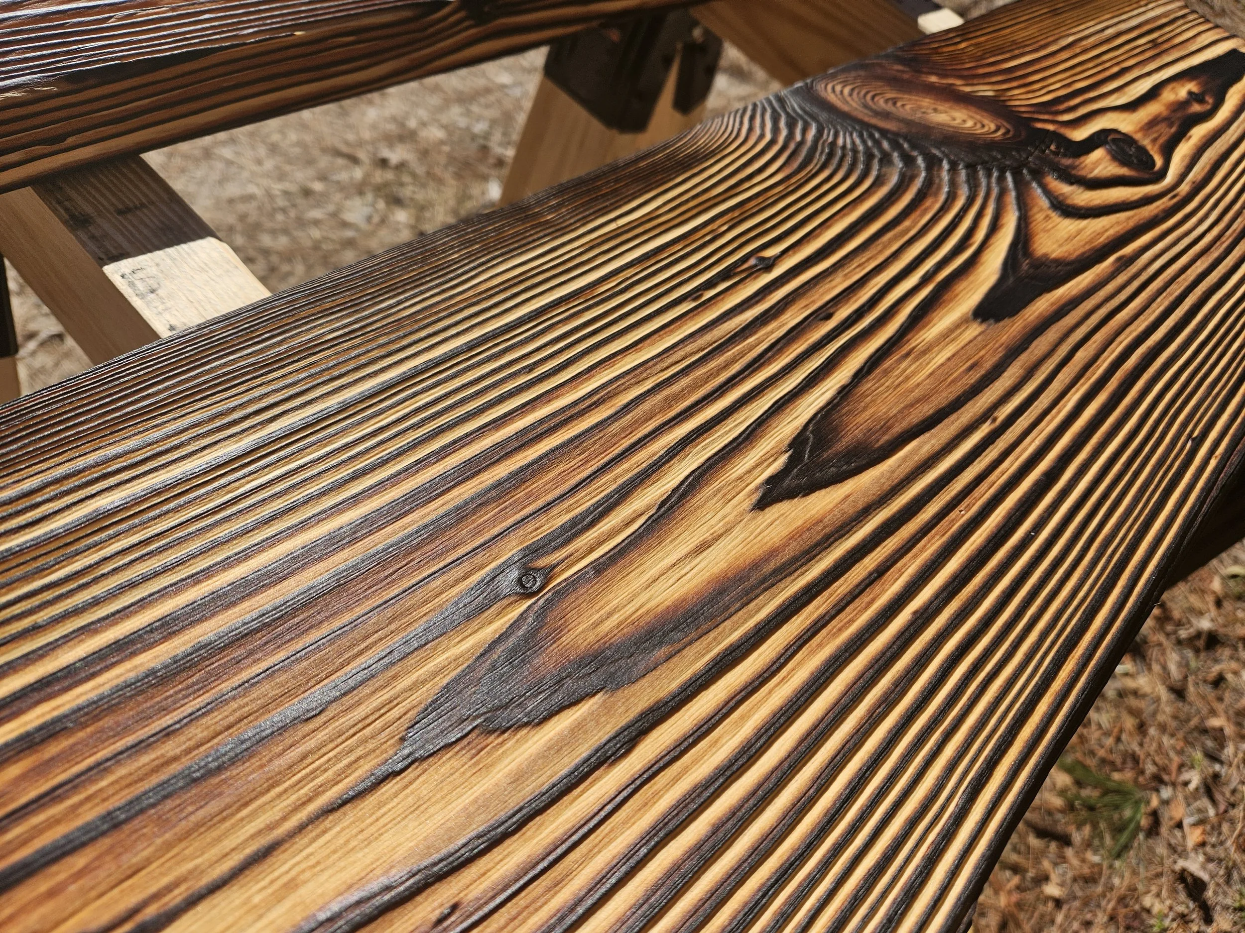 Close-up of a wooden bench with a burn mark design on the seat, outdoors on dirt ground.