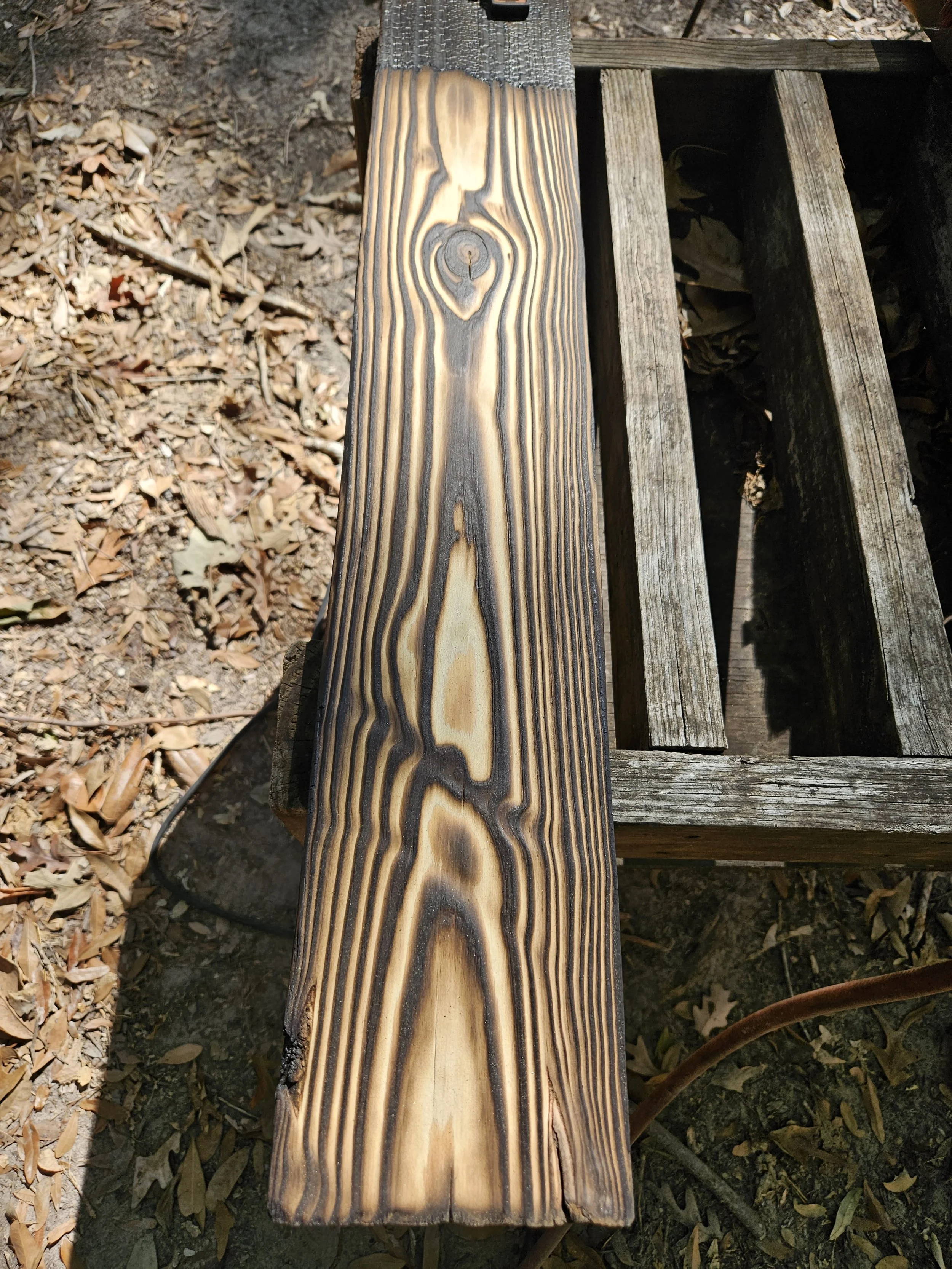 A wooden bench with a backrest showing natural wood grain and a visible knot near the top, placed outdoors on a dirt ground with dried leaves.