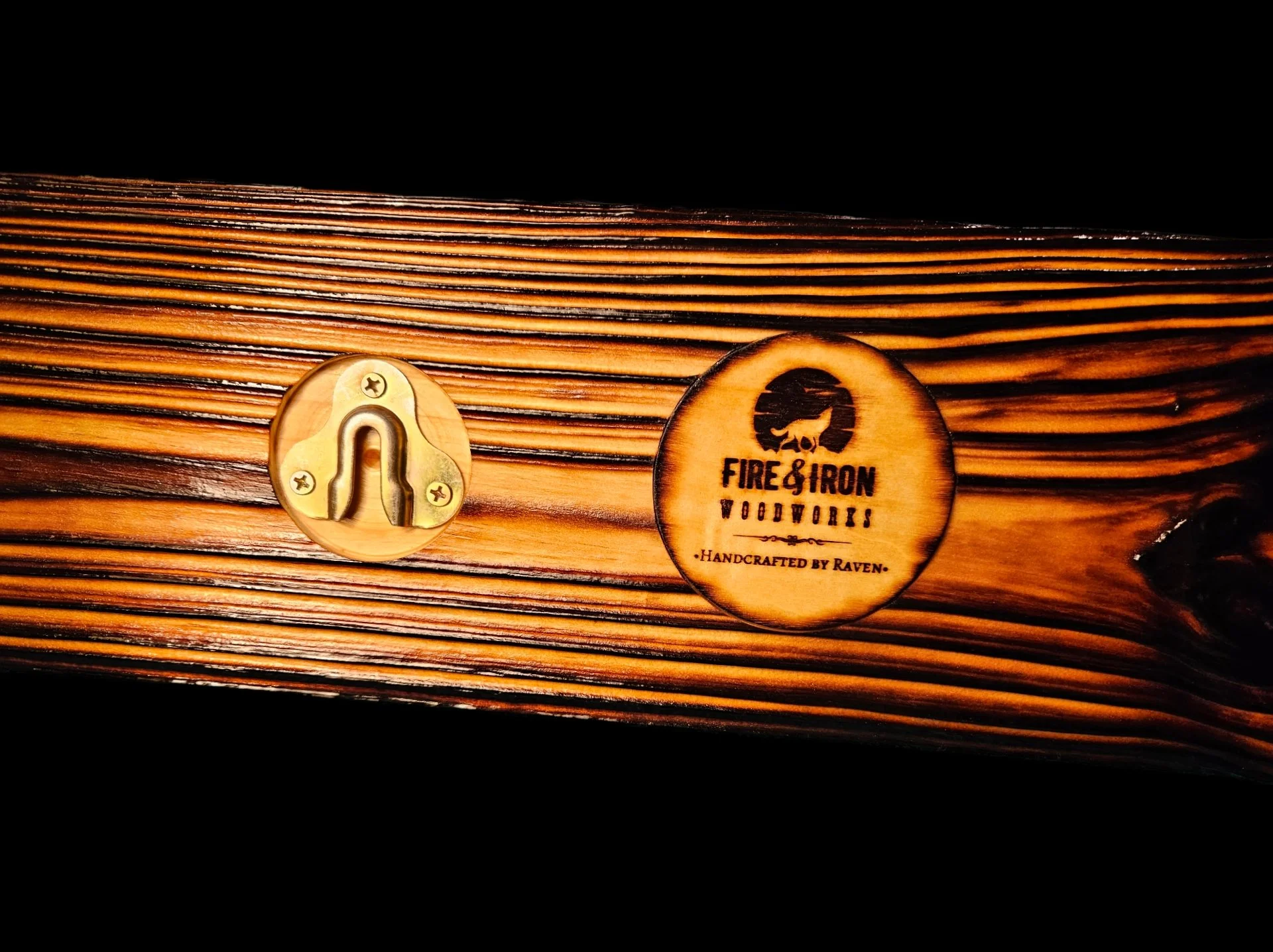 Close-up of a wooden box with the logo 'Fire & Iron Woodworks, Handcrafted by Raven,' featuring a bear silhouette, and a brass decorative piece on the side.