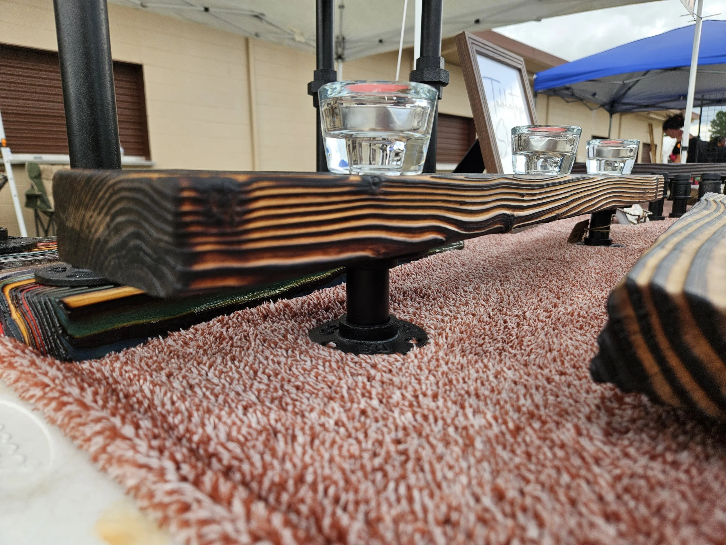Three glasses of clear liquid, possibly water, sitting on a textured wooden display stand under a canopy at an outdoor event.