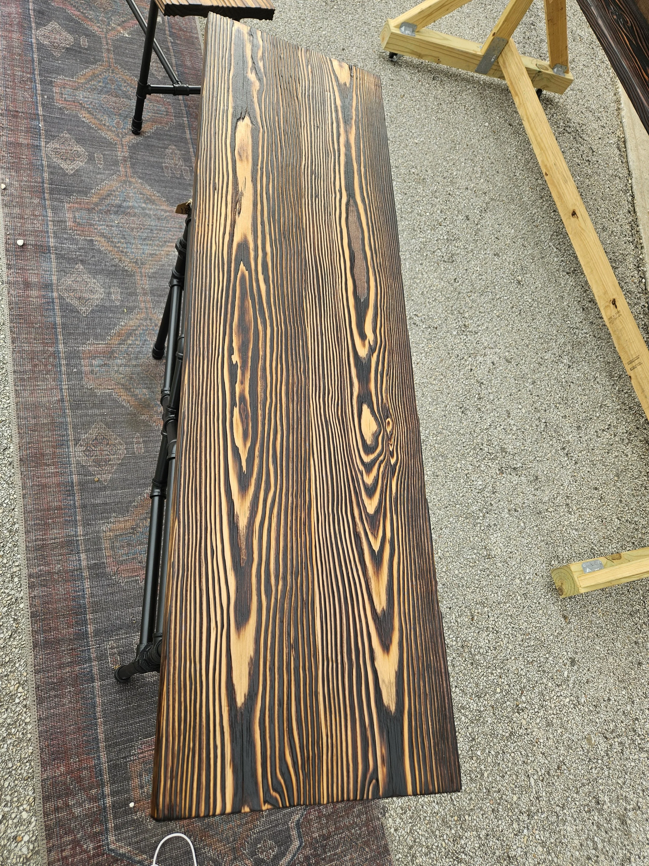 A long wooden plank with a burnt wood pattern rests on a black metal frame outdoors on a gravel surface, partially on a patterned outdoor rug.