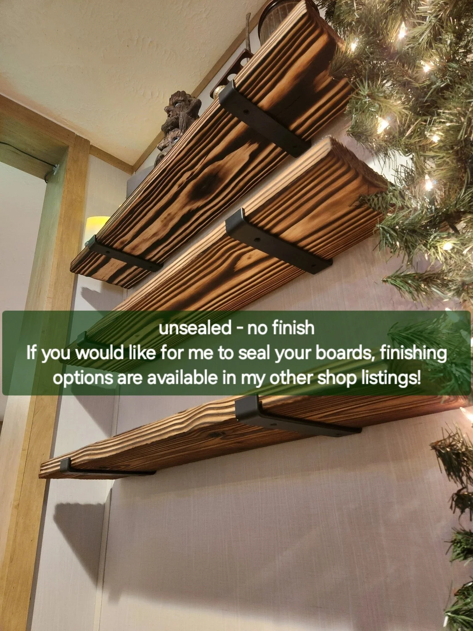 Unfinished wooden boards with dark metal brackets mounted on a white wall, partially visible Christmas tree with small lights on the right side.