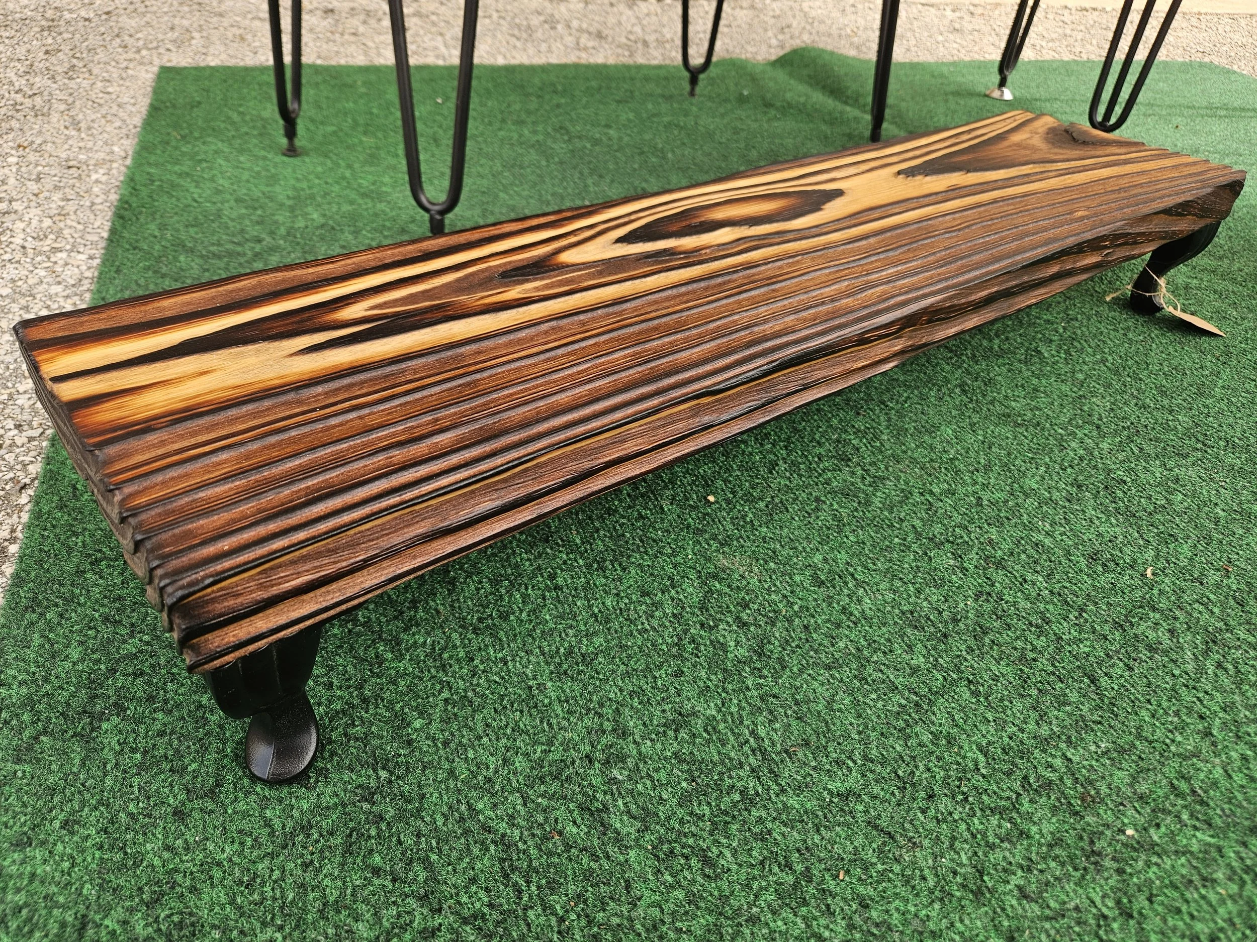 A wooden bench with dark wood grain, placed on green artificial turf, and supported by black metal legs.