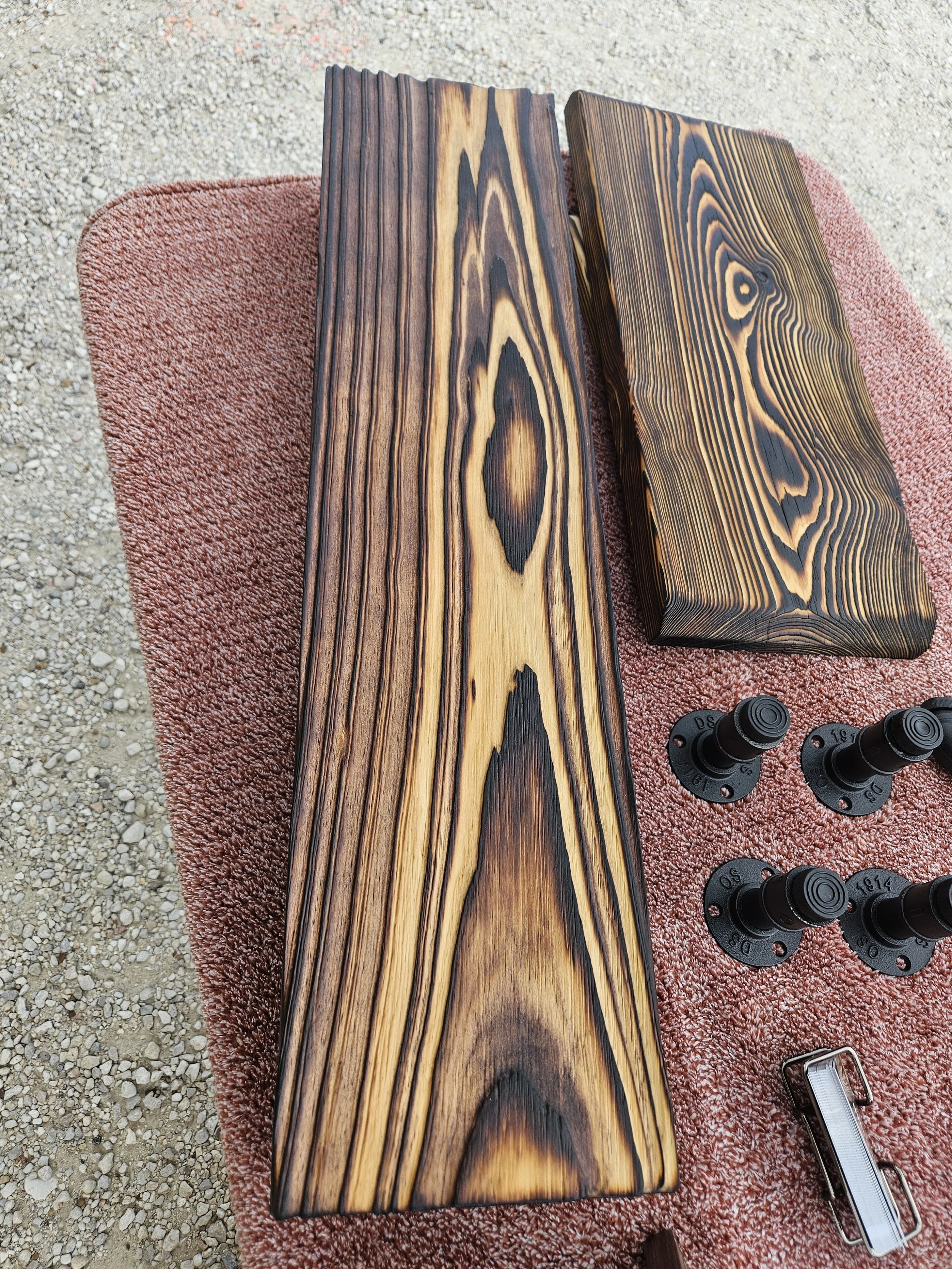 Two wooden boards with different darkened grain patterns resting on a pinkish-brown textured rug, with black camera lenses and a silver metallic object nearby, on a gravel surface.