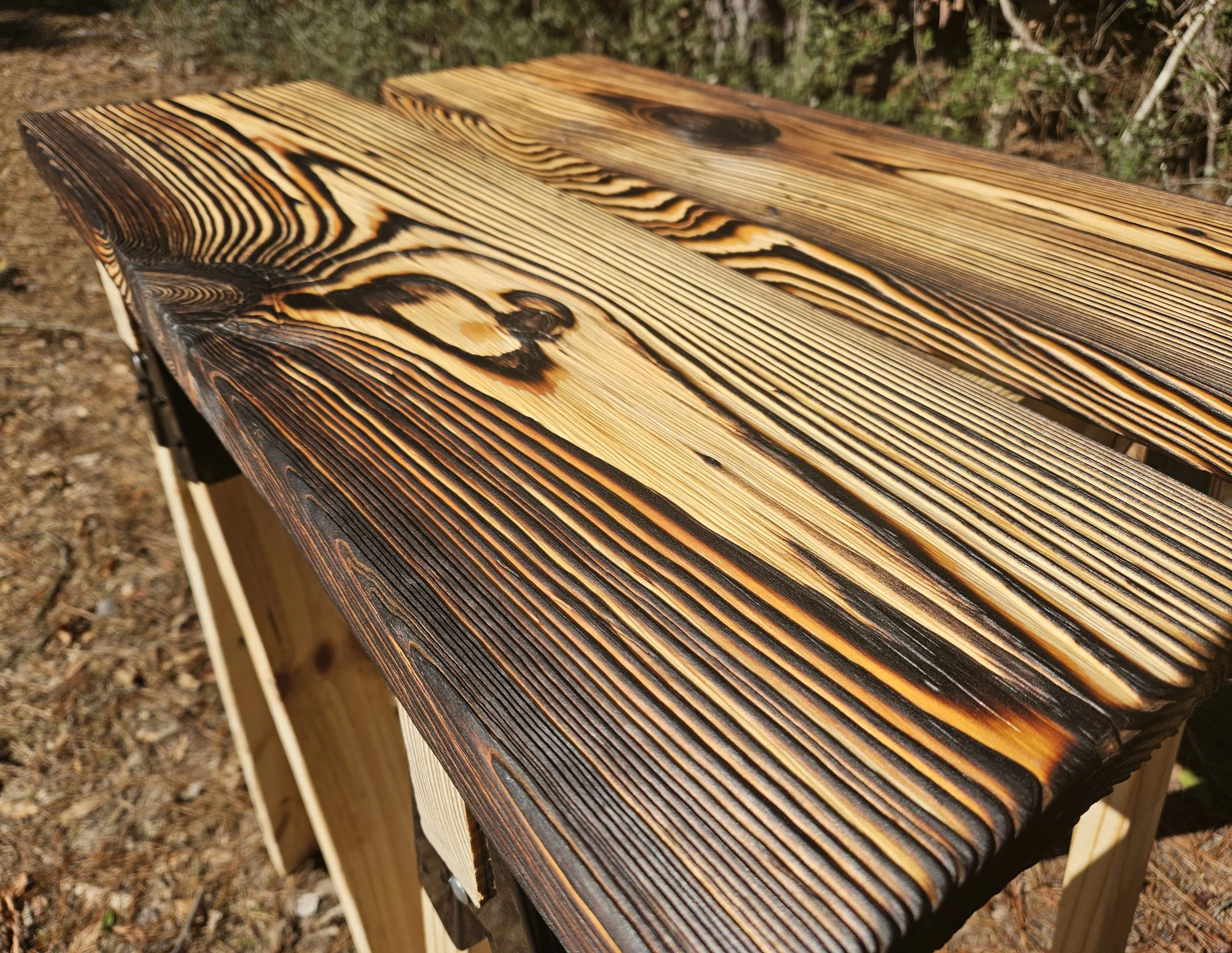 Close-up of a wooden bench with a burnt pattern on the surface, situated outdoors with pine straw and foliage in the background.