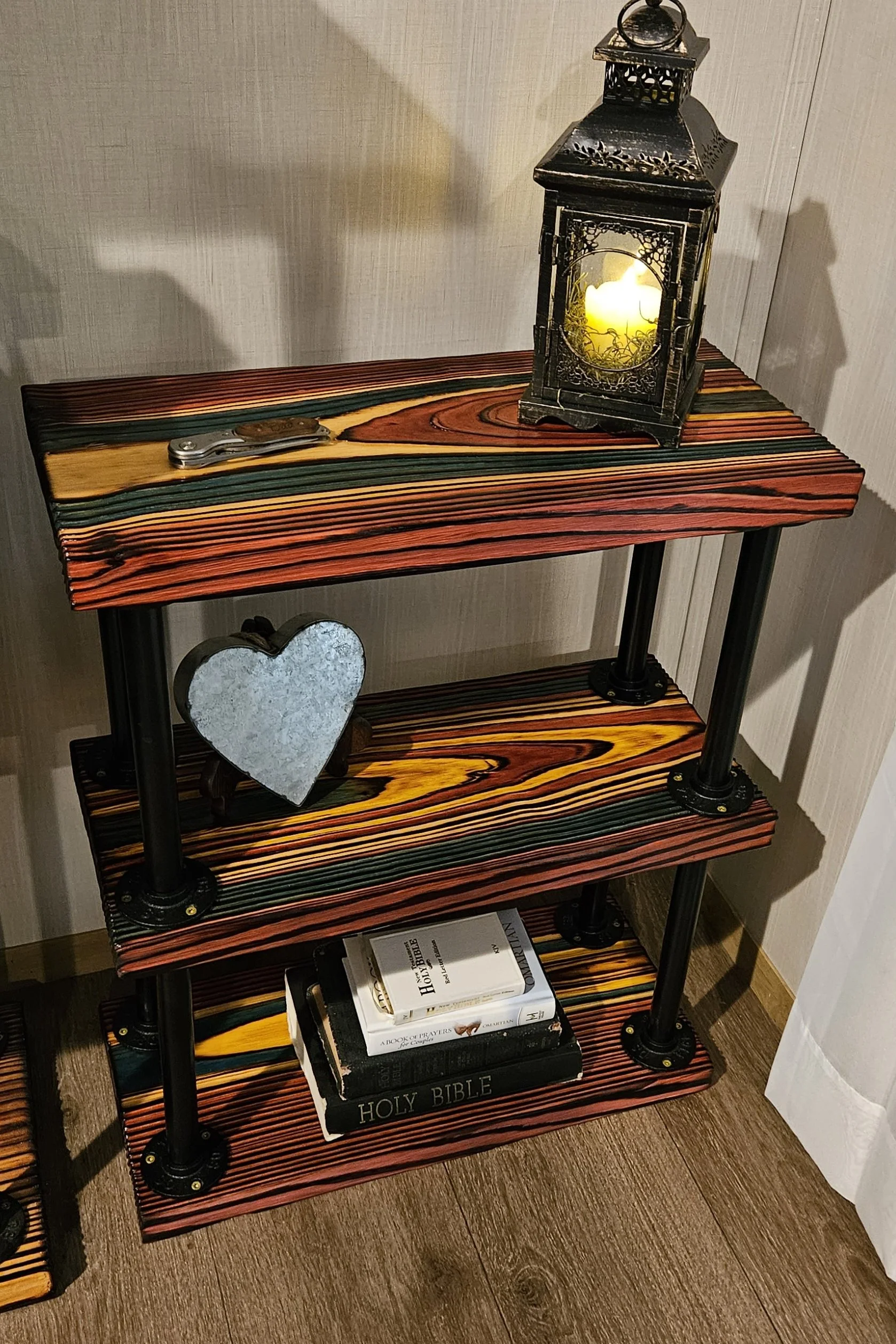 A wooden shelf with a decorative black metal lantern with a lit candle inside, a small heart-shaped mirror, and a stack of books including a Holy Bible.