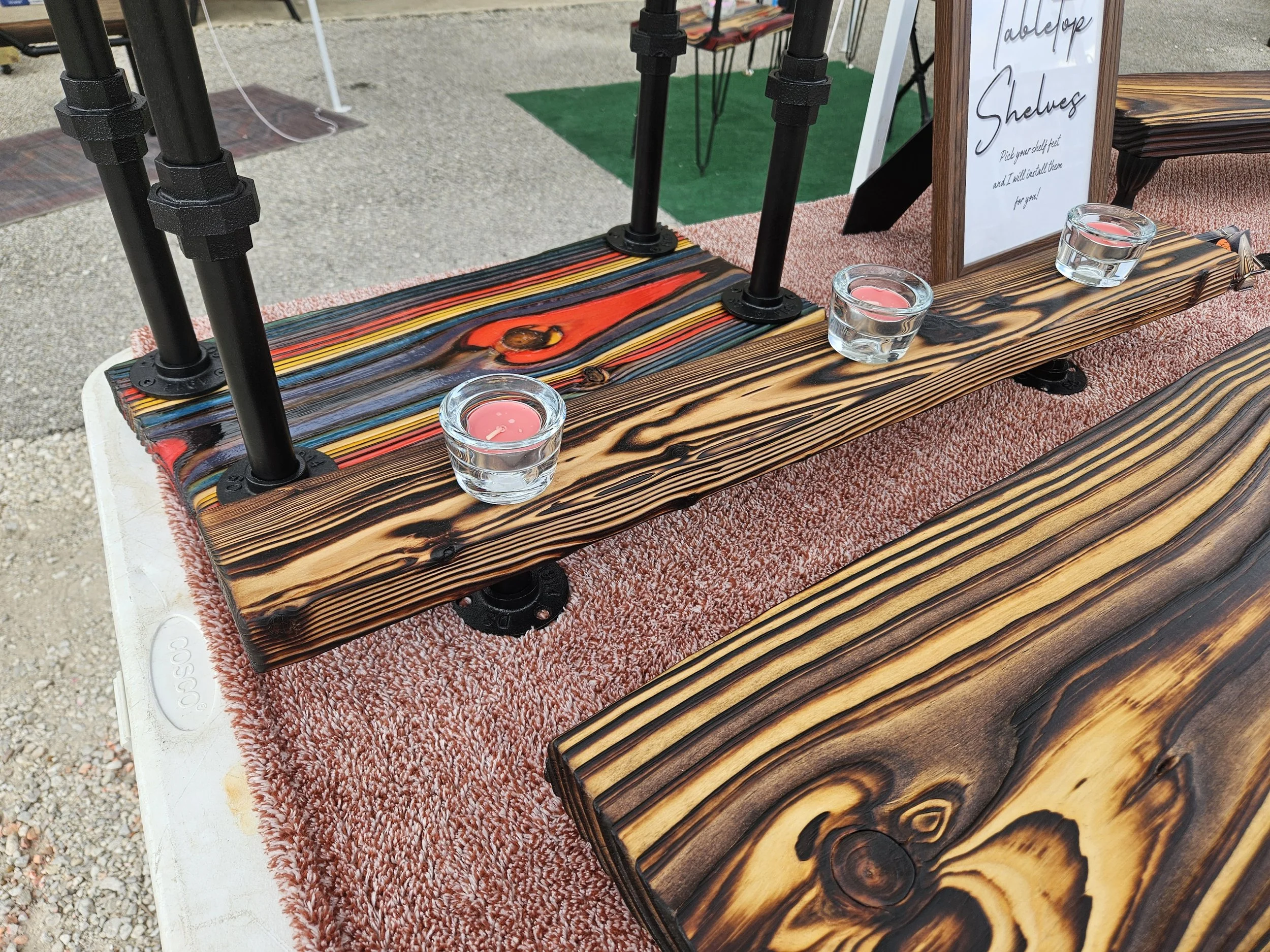 Wooden shelf with a sign that reads 'tabletop shelves' and a statement about picking shelf feet, with small glass cups holding tealight candles, on a pink textured surface at an outdoor market or fair.