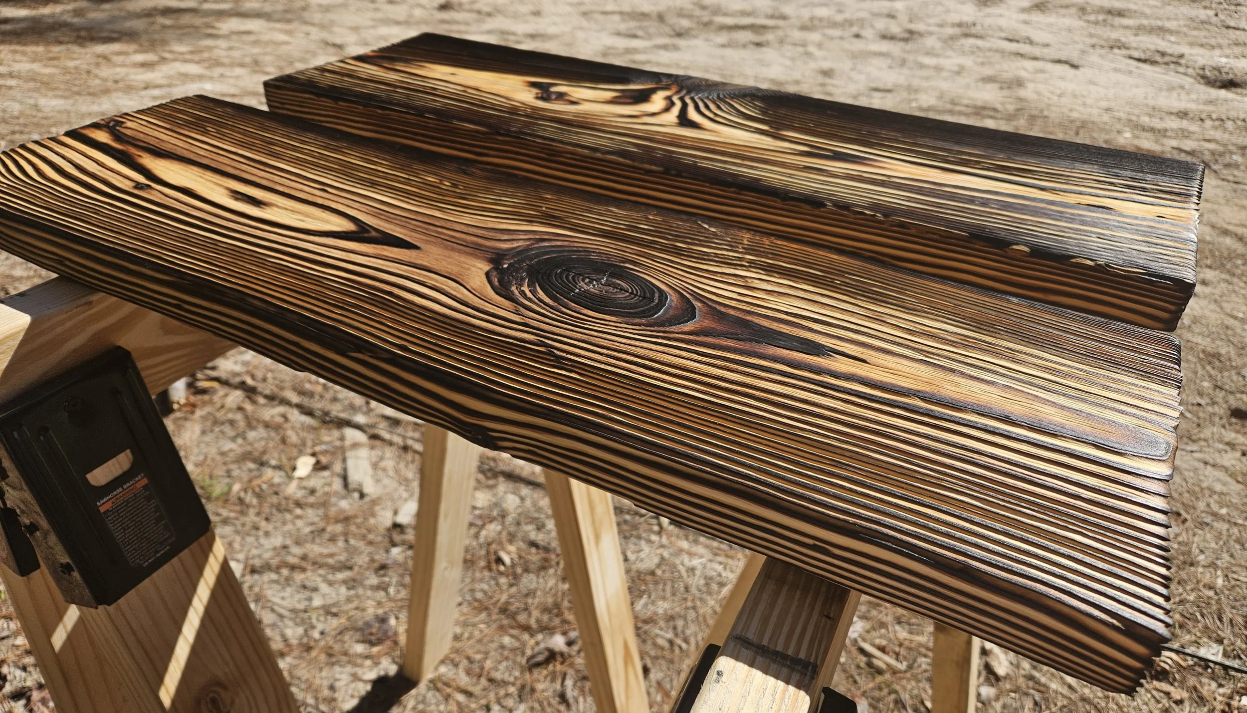 Close-up of a wooden plank with a burned finish, showing wood grain and knots, placed on a sawhorse outdoors.