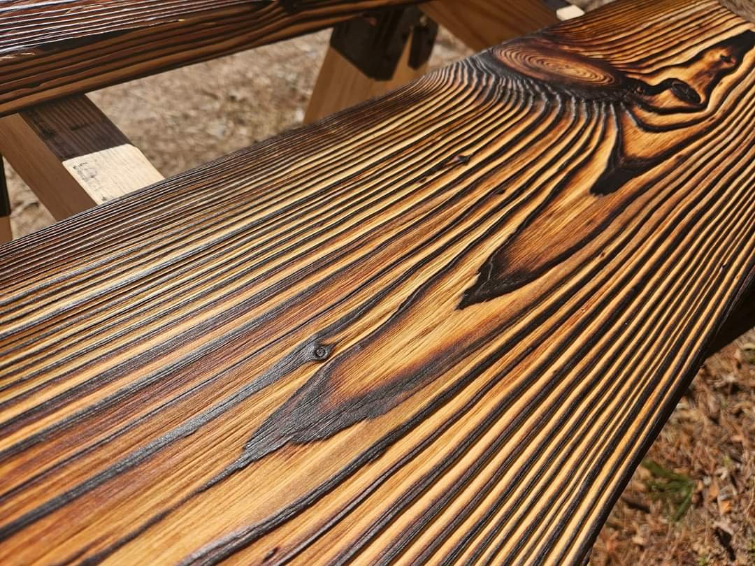 Close-up of a wooden outdoor bench with a natural finish, showing detailed wood grain and knots, set on a surface with dried leaves.