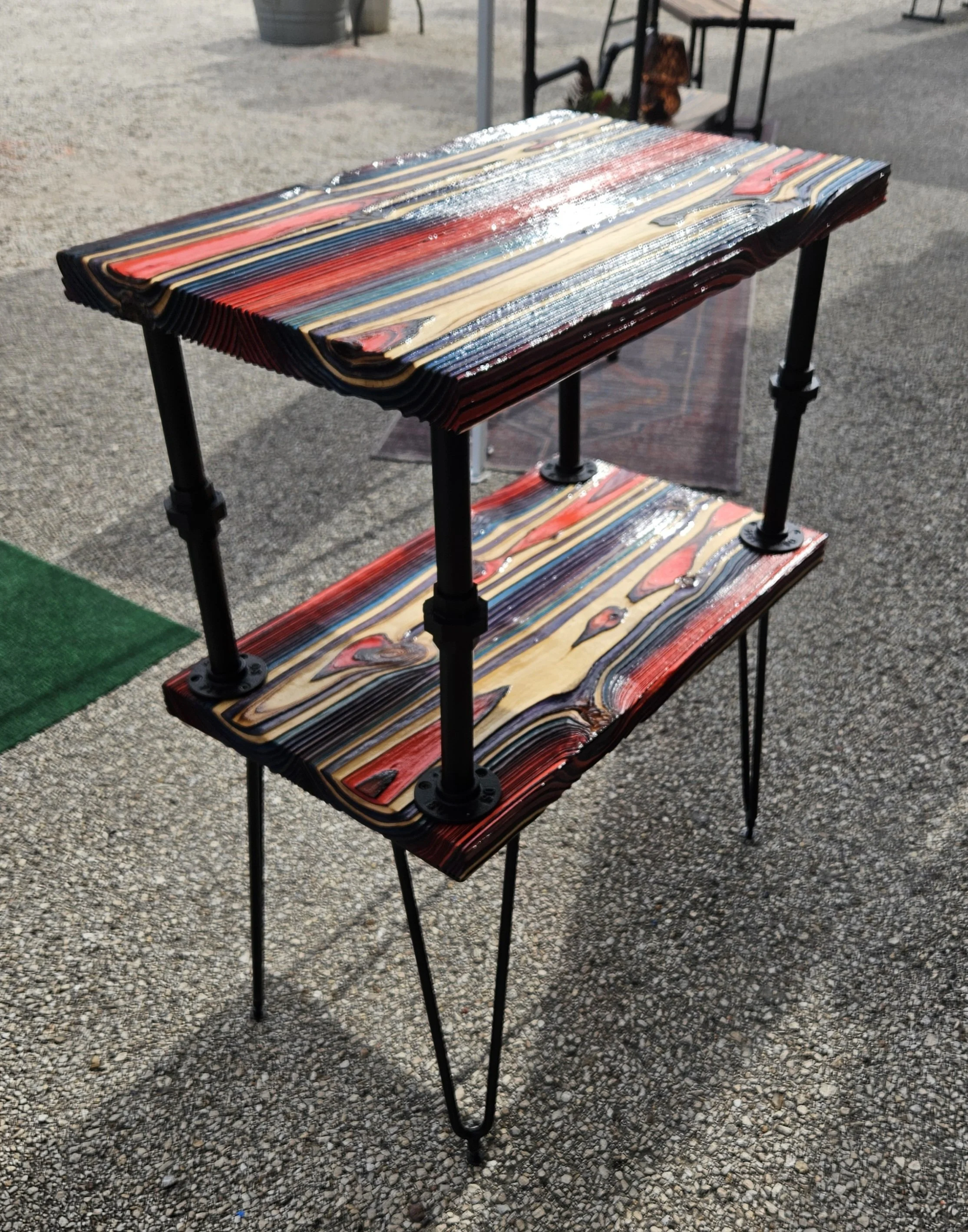 A colorful, striped wooden table with metal legs outdoors on pavement, with another table and chairs visible in the background.