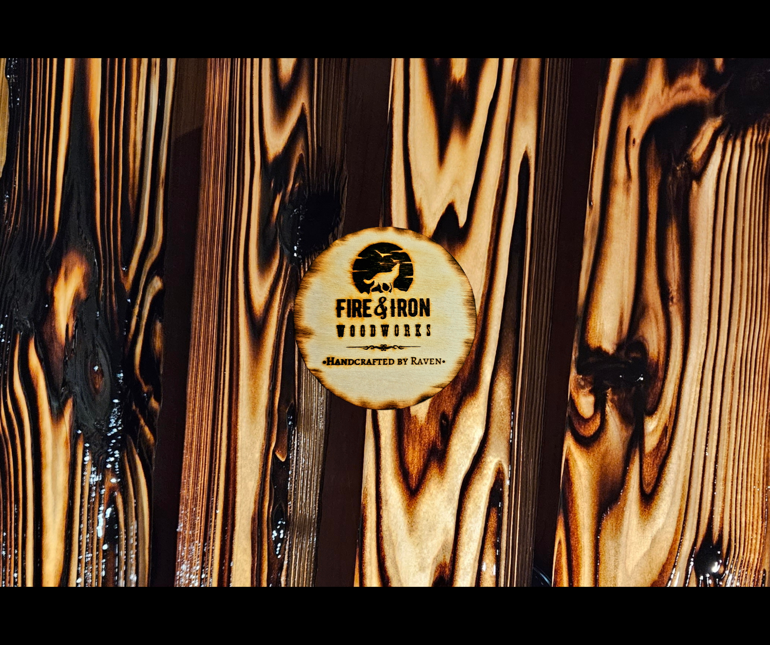 Wooden surface with a round logo that reads "Fire & Iron Woodworkers, Handcrafted by Raven" featuring a silhouette of a wolf.
