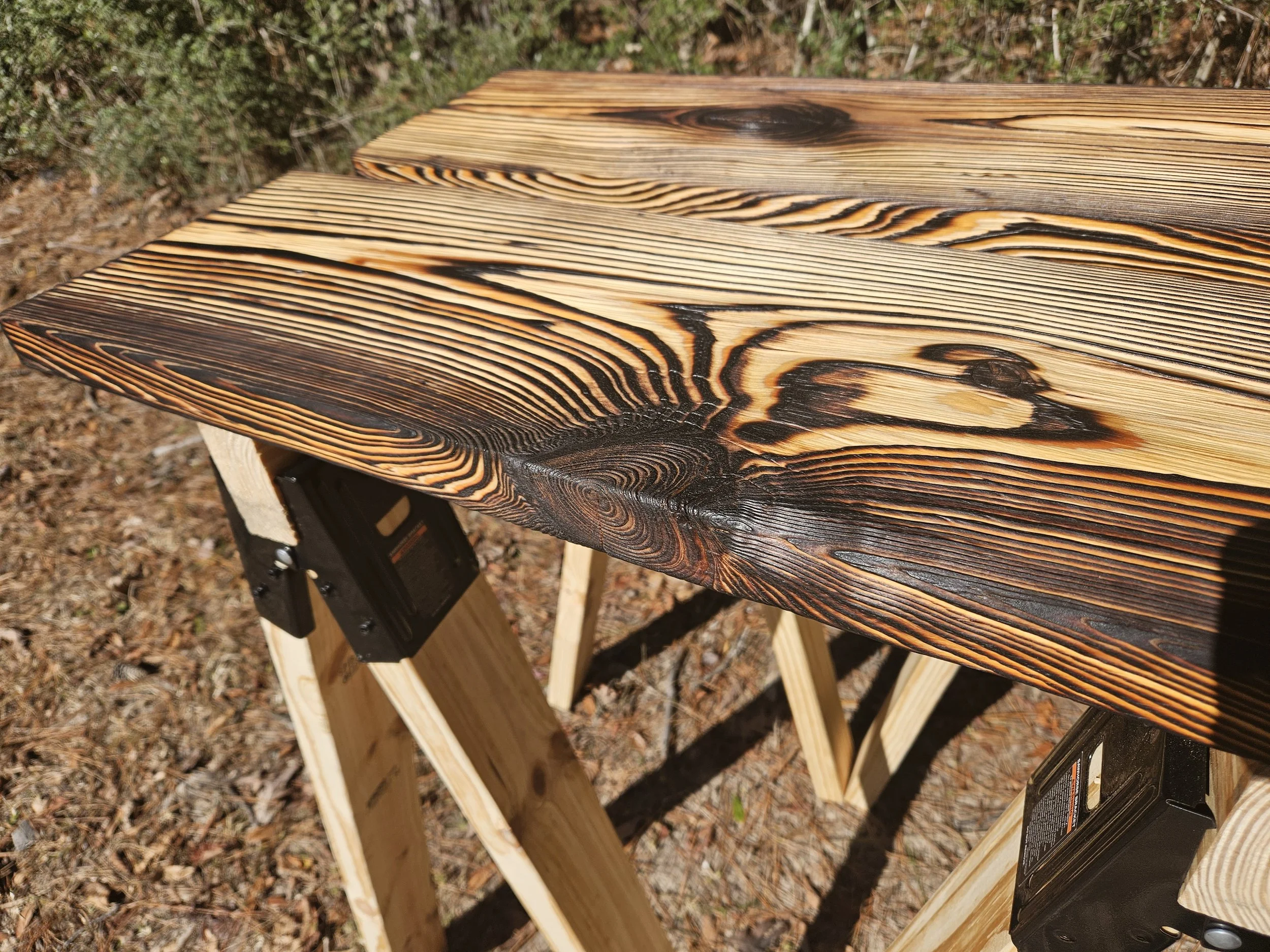 Close-up of a wooden bench with a natural wood finish, showing the detailed grain and knots, outdoors on a bed of fall leaves and pine needles.