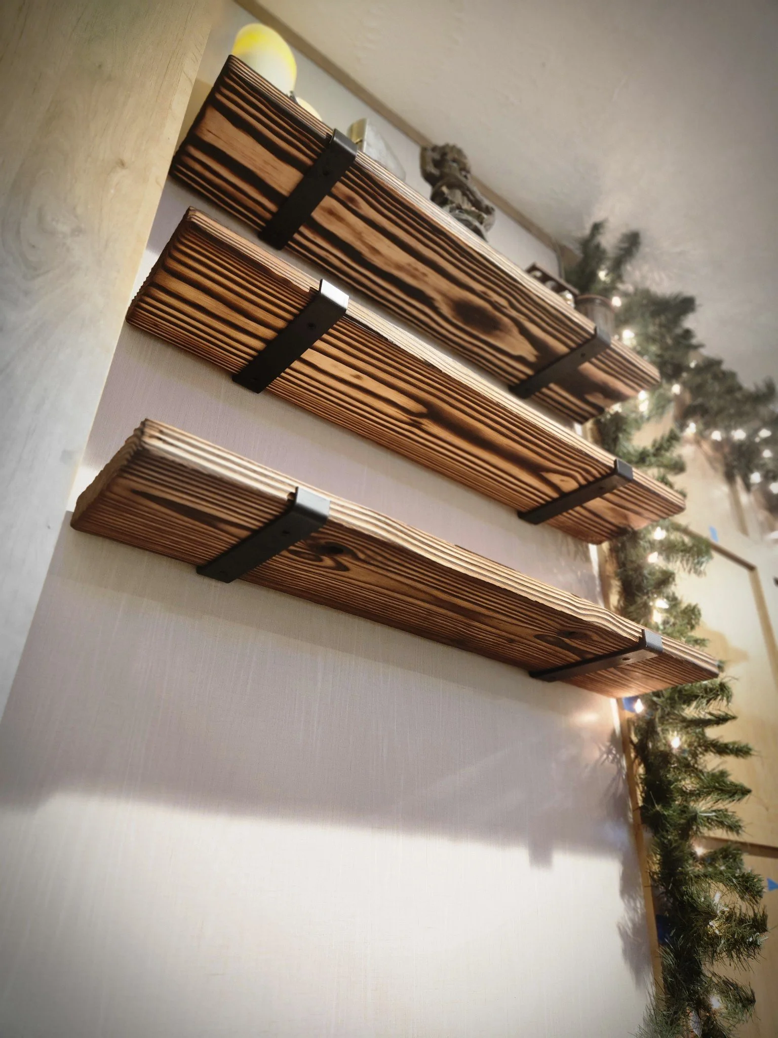 Three wooden shelves with black brackets mounted on a white wall, with Christmas greenery and lights in the background.