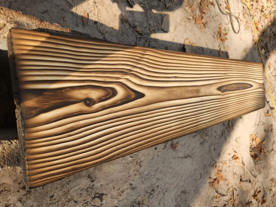 A close-up of a wooden board with visible grain patterns and knots, resting on the ground outdoors.