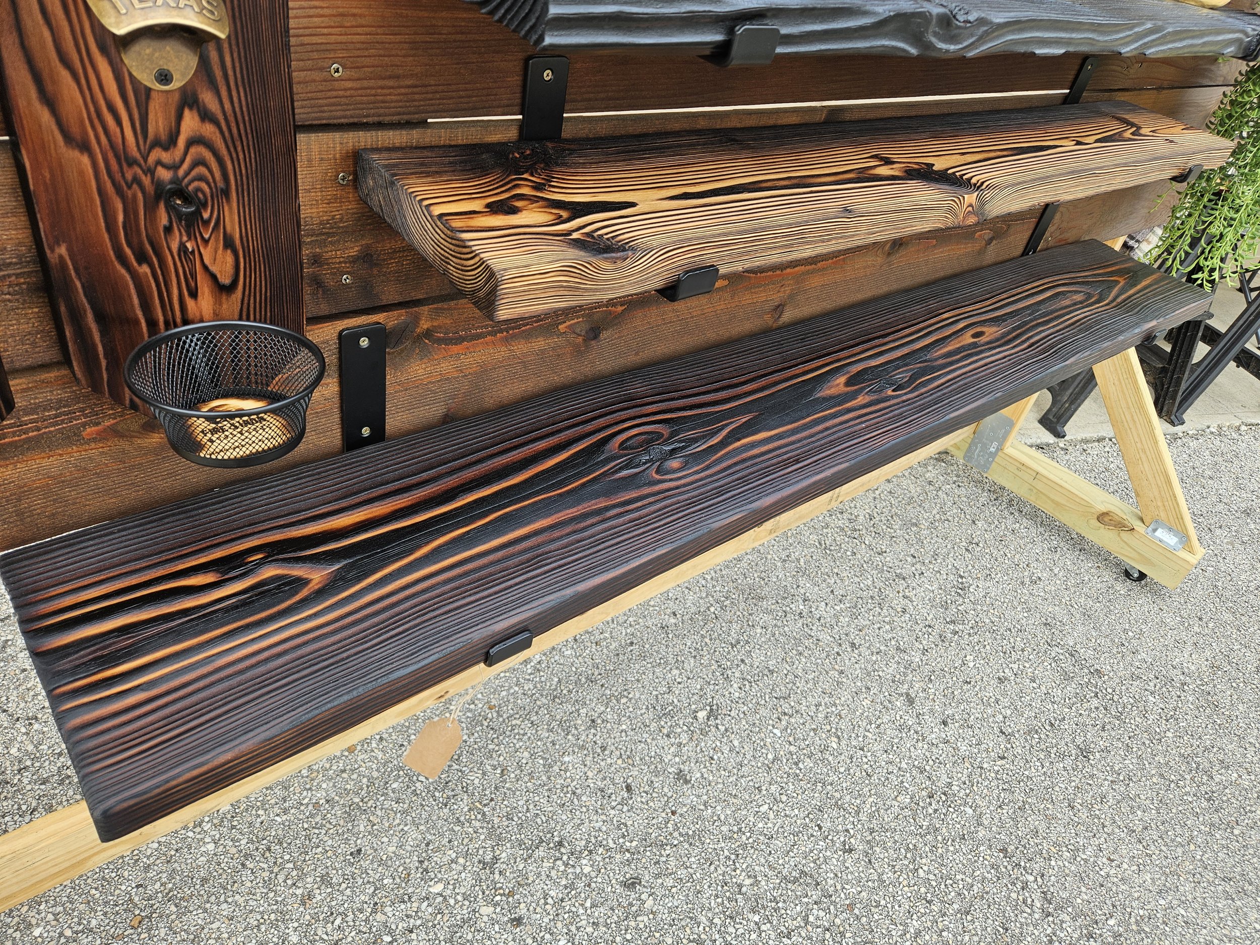 Wooden outdoor benches with black metal supports, arranged in rows. There is a small black wire mesh trash bin attached to the side of one bench. The benches are placed on gravel ground and there is some greenery visible on the right side of the imag