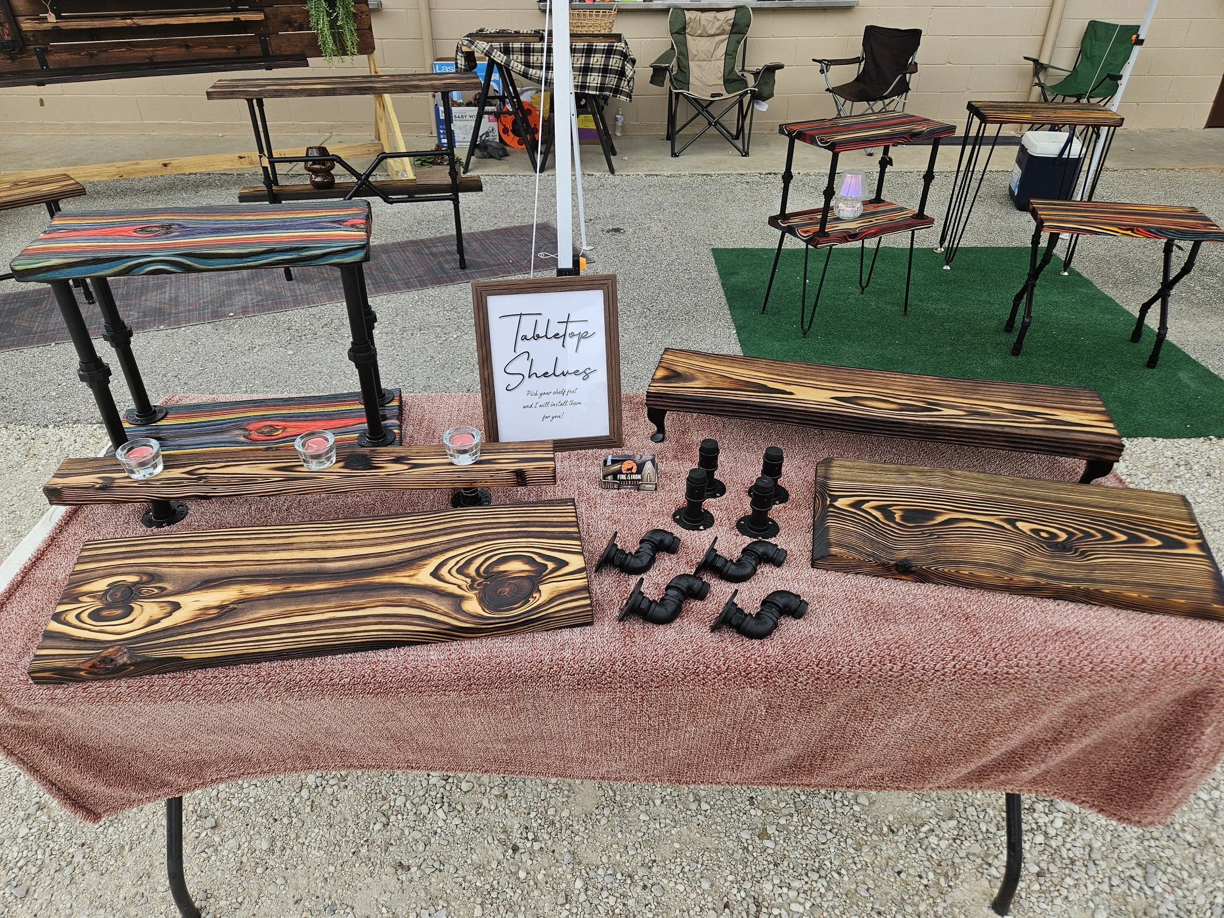 Outdoor display of wooden tabletop shelves and small tables with striped and natural wood finishes, set on a gravel surface with a small green rug, including a sign that reads 'Tablentope Shelves' and some small candles and bottle decorations.