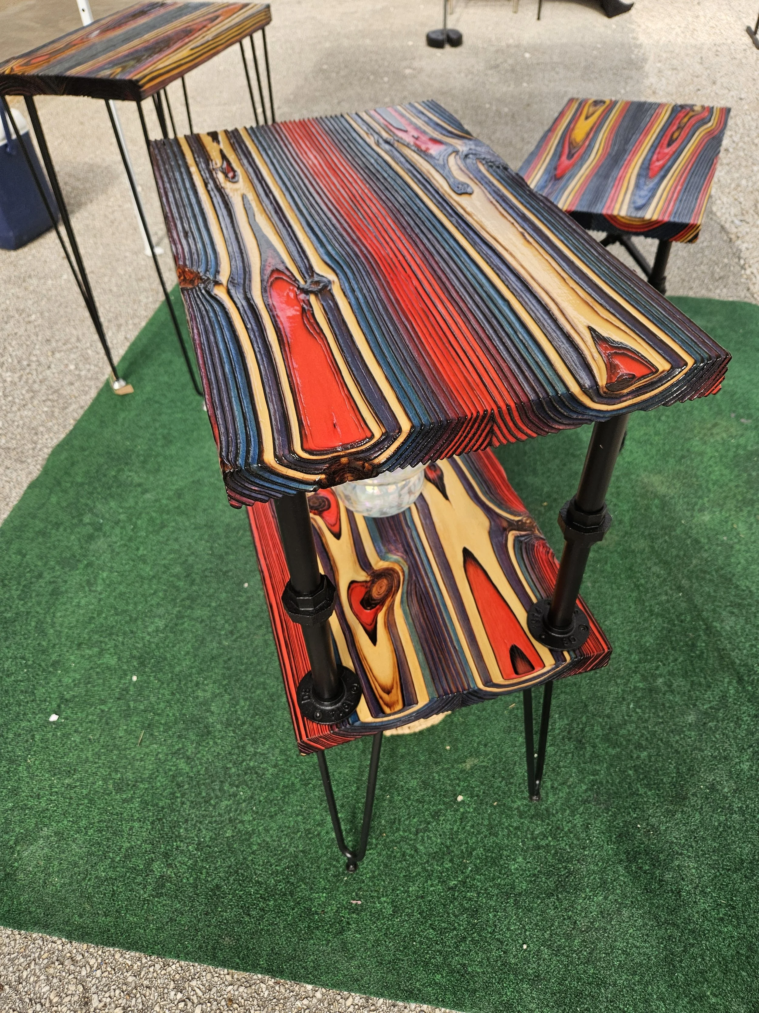 Colorful, layered wooden tables with a striped pattern in red, yellow, blue, black, and natural wood tones, displayed on black metal stands on green artificial turf with gravel surrounding.