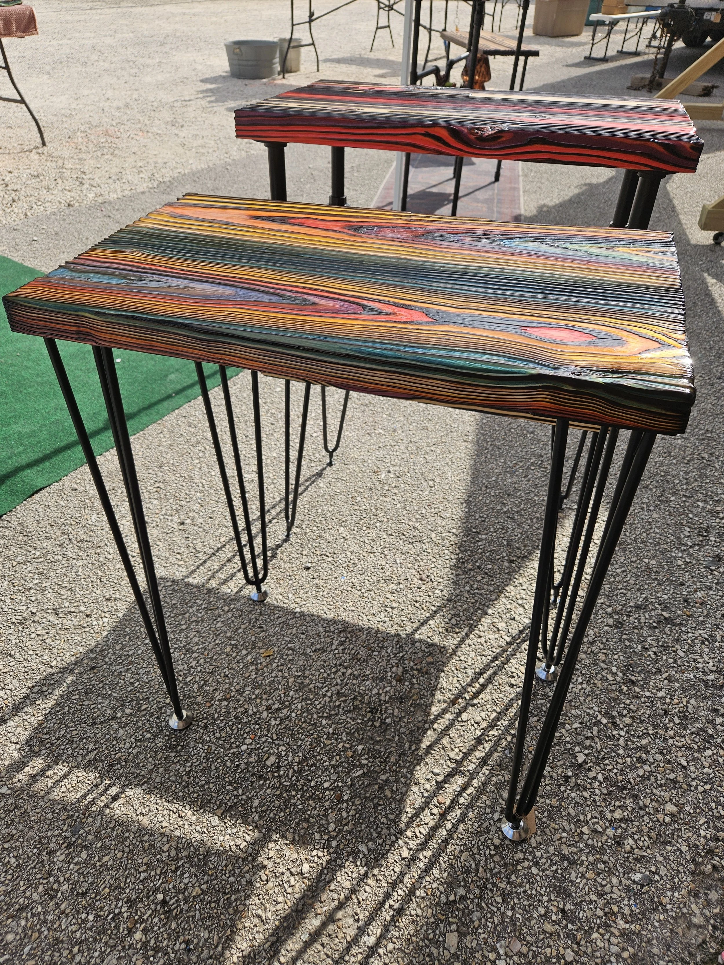 Two wooden tables with black metal hairpin legs outdoors on a gravel surface, with various outdoor furniture and objects in the background.
