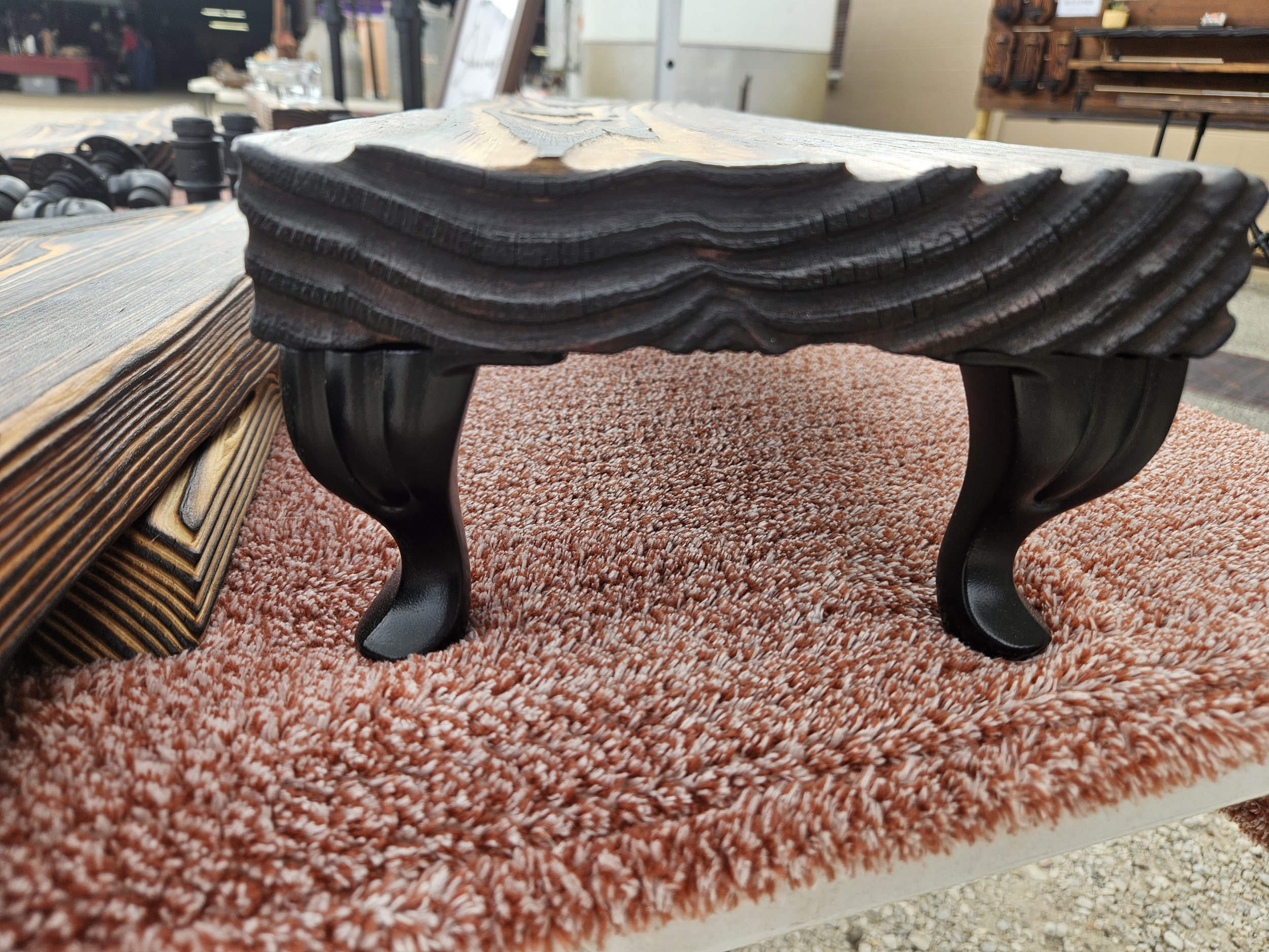 Close-up of a wooden coffee table with a black, ornate base, placed on a pink and white patterned carpet, with other furniture and store shelves in the background.