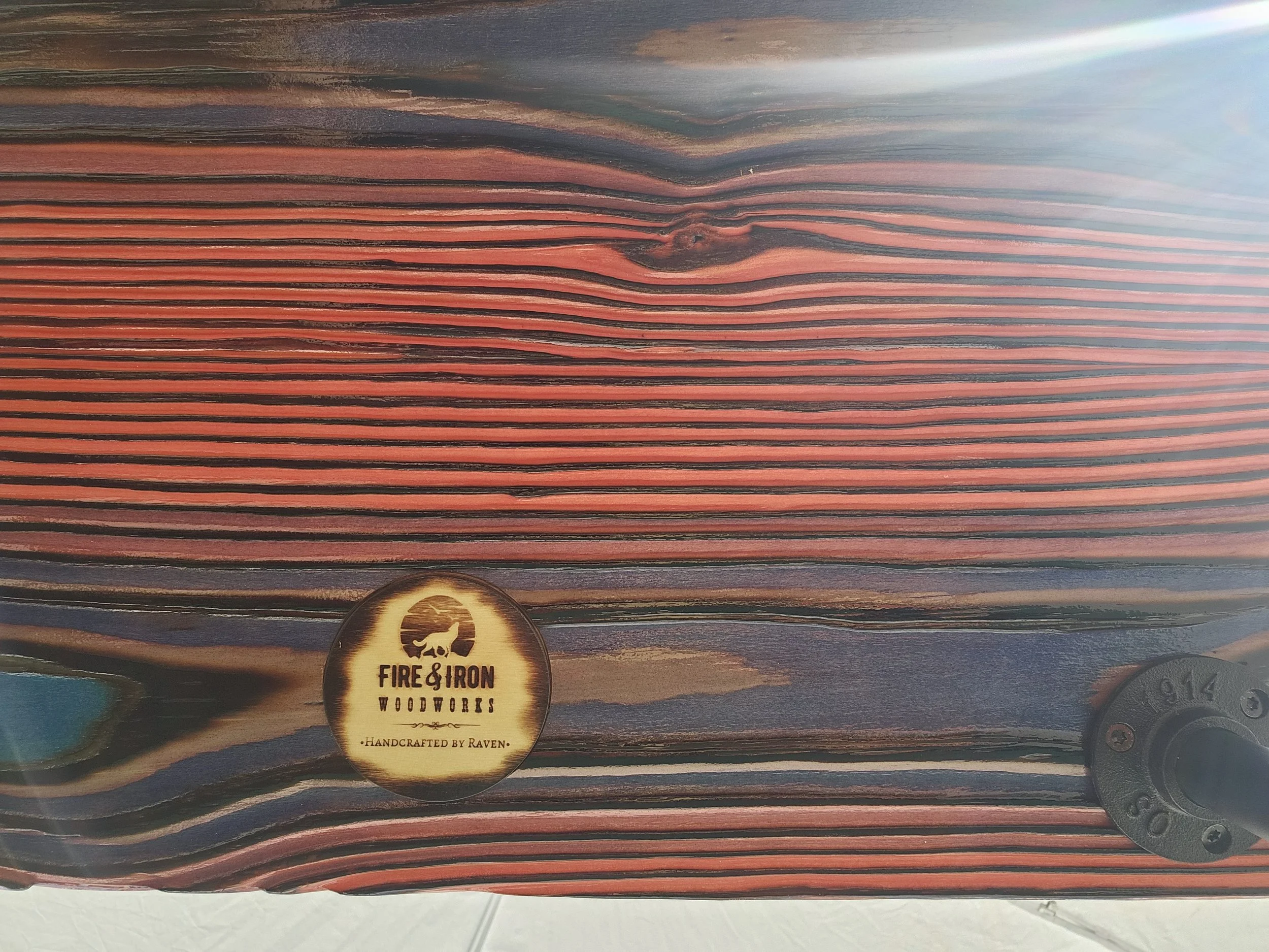 Close-up of a woodgrain surface with a logo at the bottom center that reads 'Fire & Iron Woodworks - Handcrafted by Raven' with an image of a wolf.