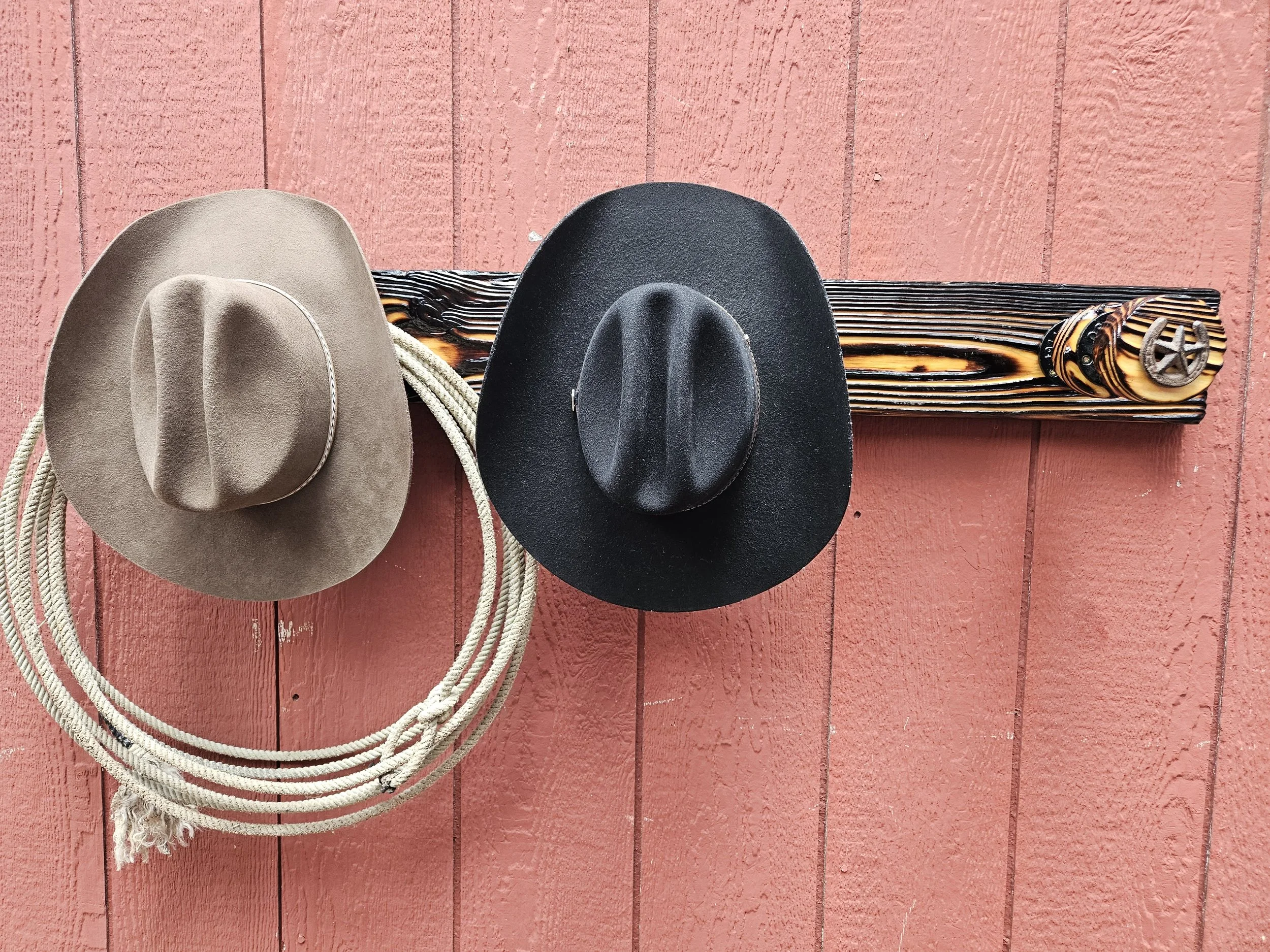 A beige cowboy hat and a black wide-brimmed hat resting on a rustic, striped wooden post with decorative metal accents, against a red wooden wall background.
