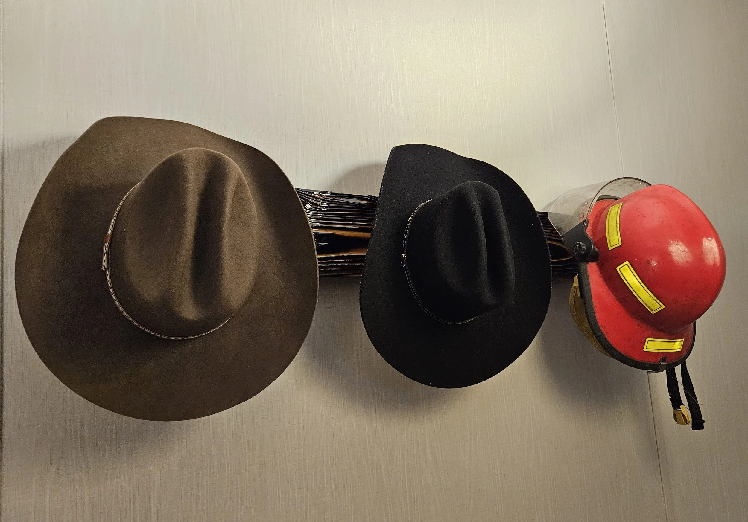 Two cowboy hats, one brown and one black, are hanging on a metal rack. There is a red firefighter helmet with a yellow reflective stripe and a clear plastic visor hanging on the same rack. Behind the rack are some black file folders stacked verticall