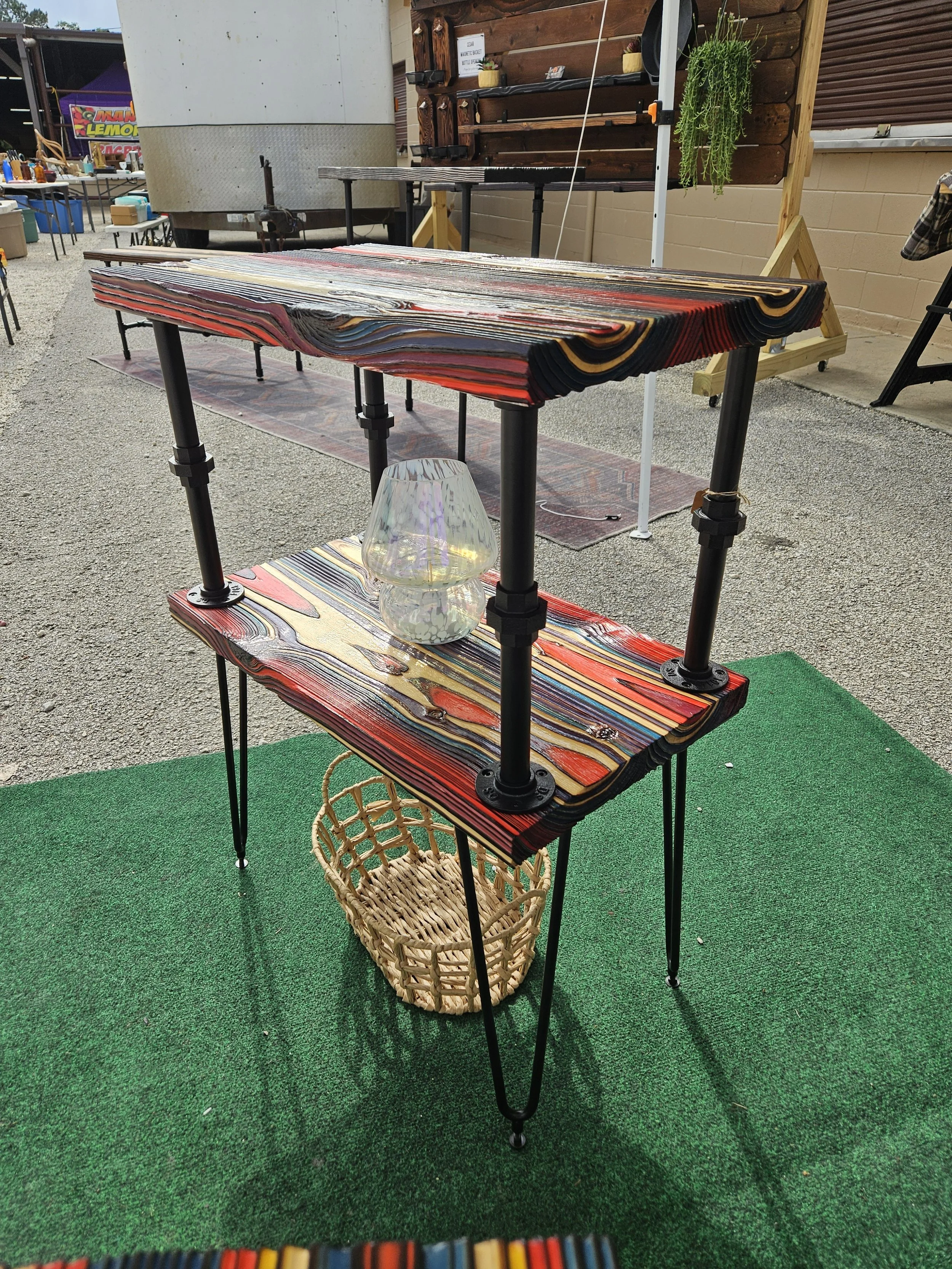 A display of colorful layered wood pieces arranged on a three-tier black metal stand with a small wicker basket underneath. The setup is outdoors on green artificial turf.