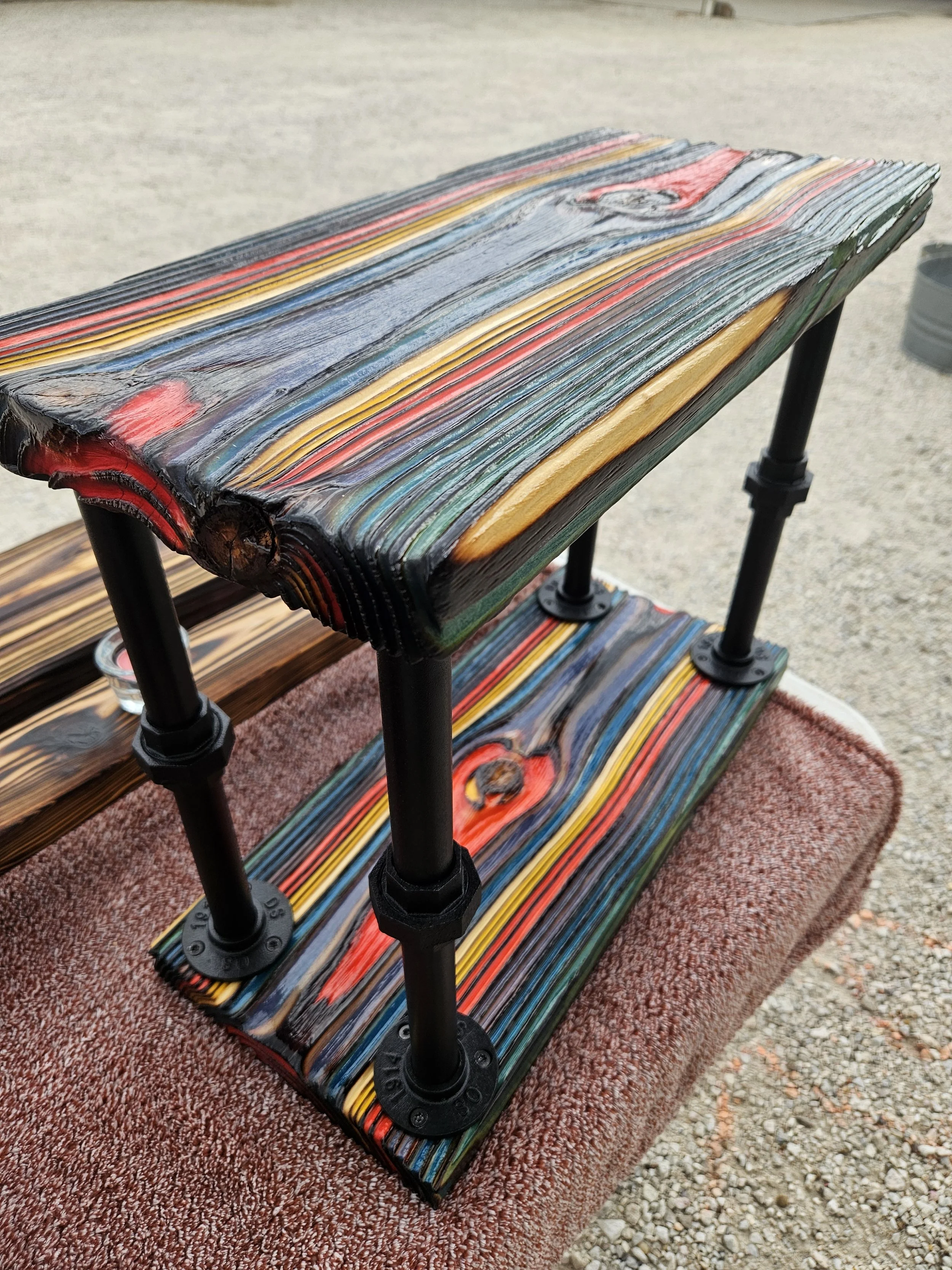 A multi-tiered wooden shelf with colorful, painted wood planks, supported by black metal pipes, placed on a red carpet outdoors.