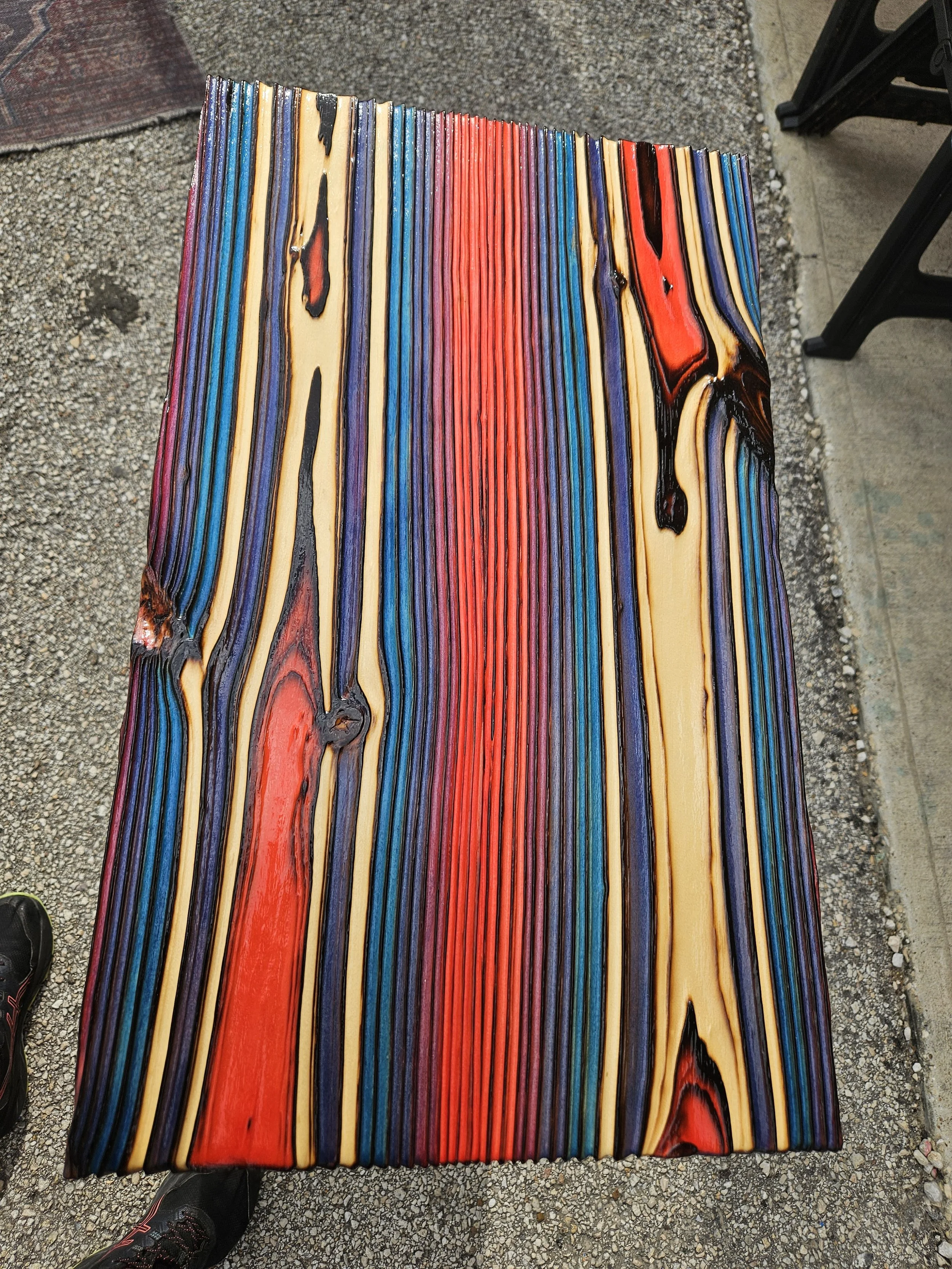 A wooden bench with multicolored, swirl-patterned paint, situated on a gravel surface outdoors.