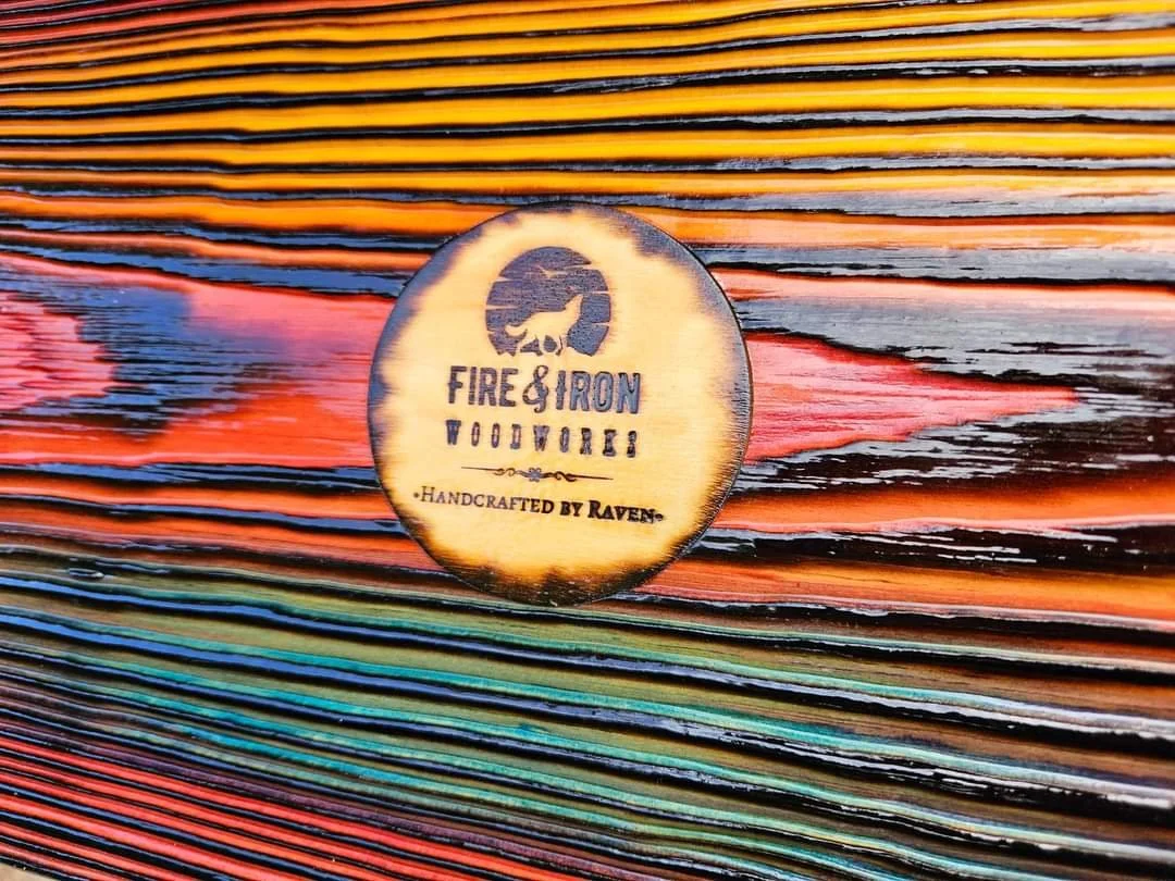A wooden coin with the logo and text for Fire & Iron Woodworks, handcrafted by Raven, placed on a multicolored wooden surface.