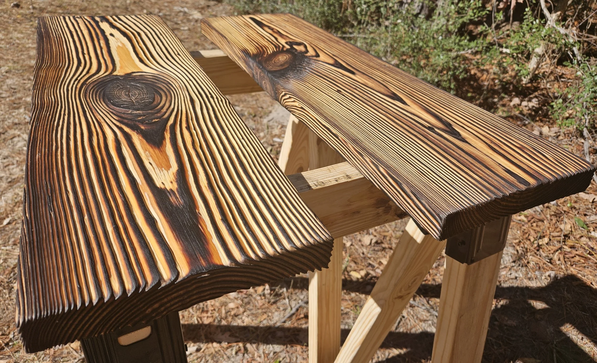 Two wooden benches with charred wood tops outdoors on dirt ground with bushes in background.