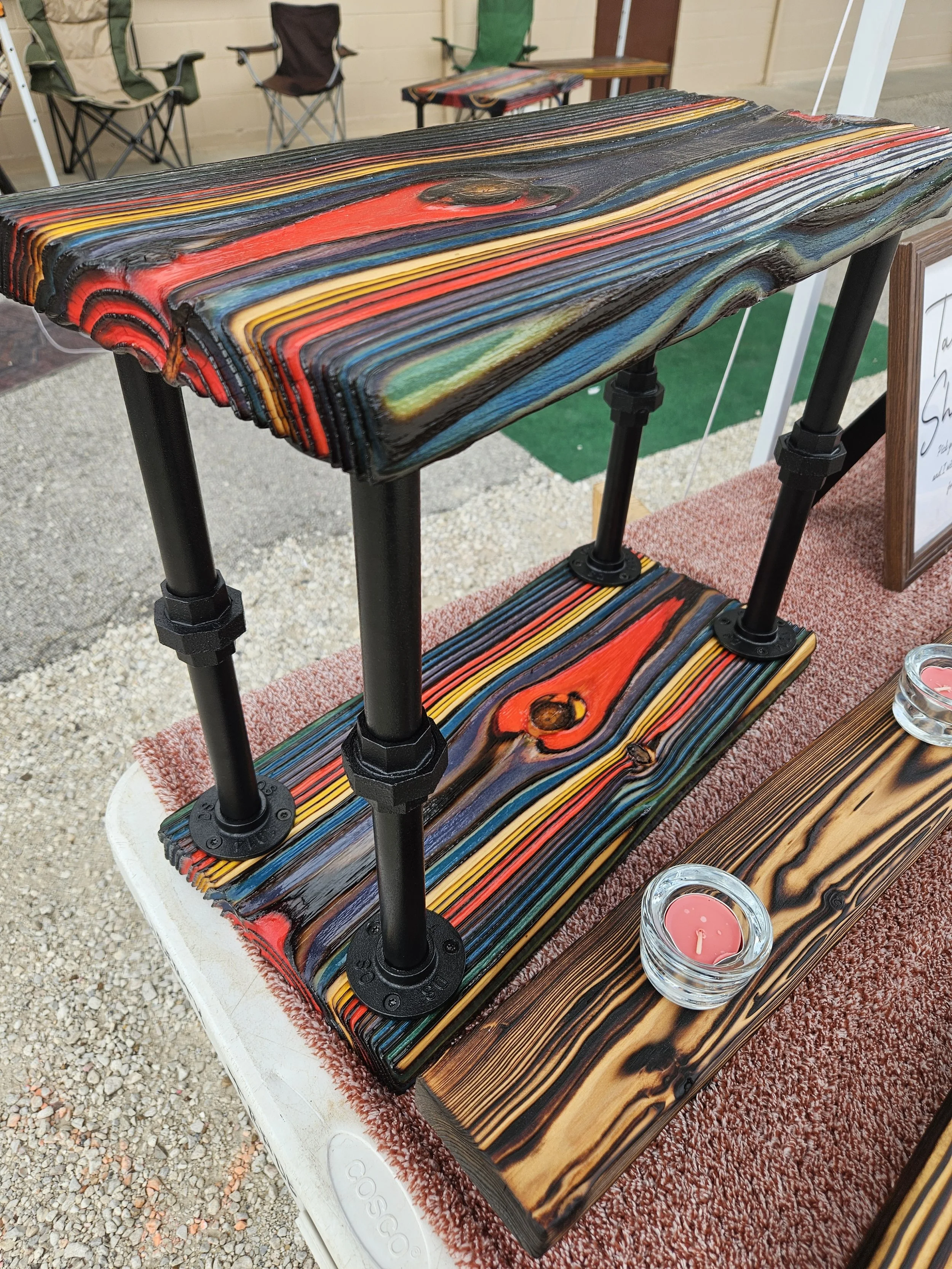 Colorful wooden tables with black metal legs displayed at an outdoor market or craft fair, with pink candles in glass holders nearby.