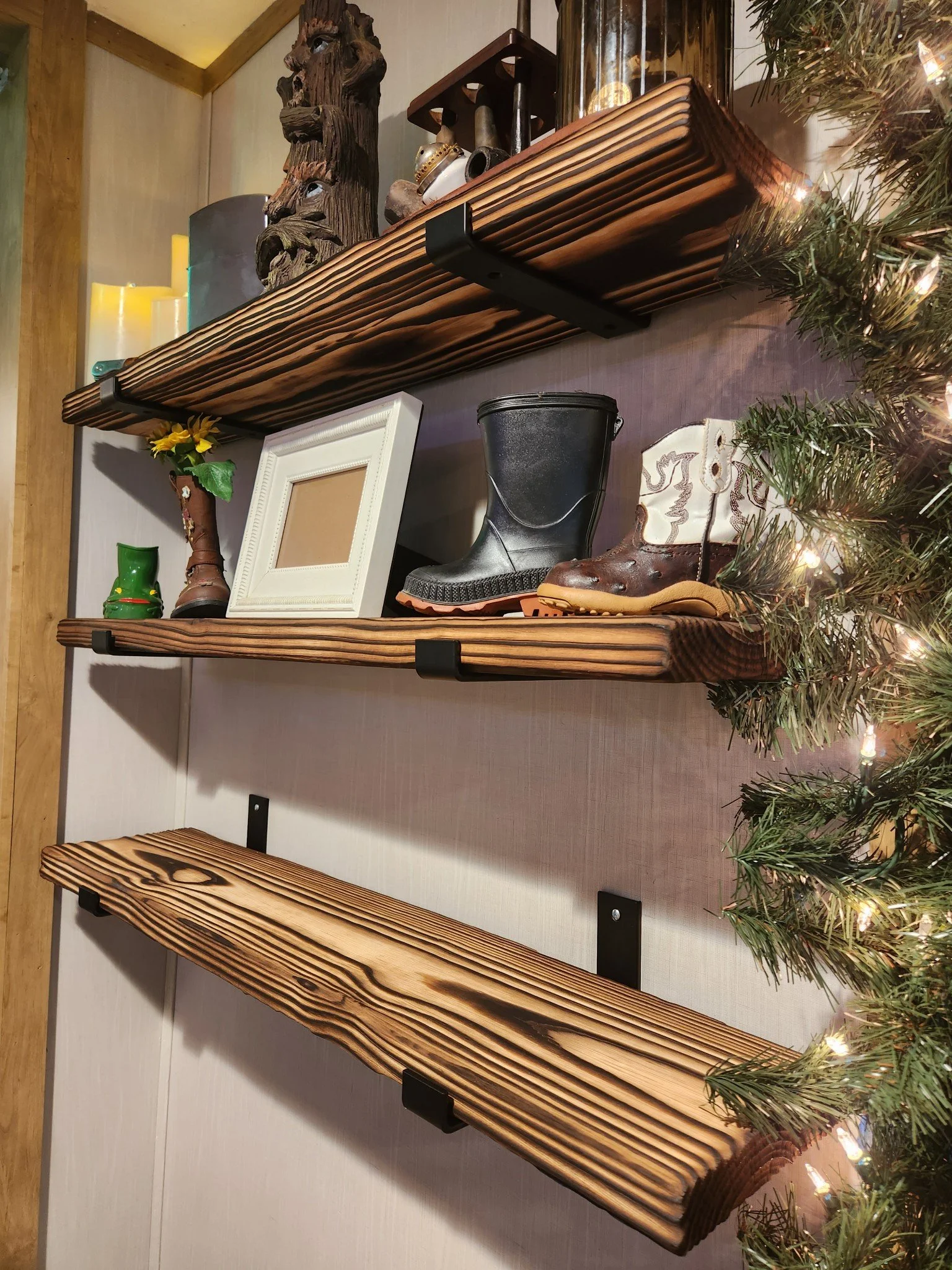Wooden shelves with various decorative items, including a carved wooden face sculpture, a white picture frame, a pair of boots, a small plant, candles, and holiday lights, with a Christmas tree to the right.