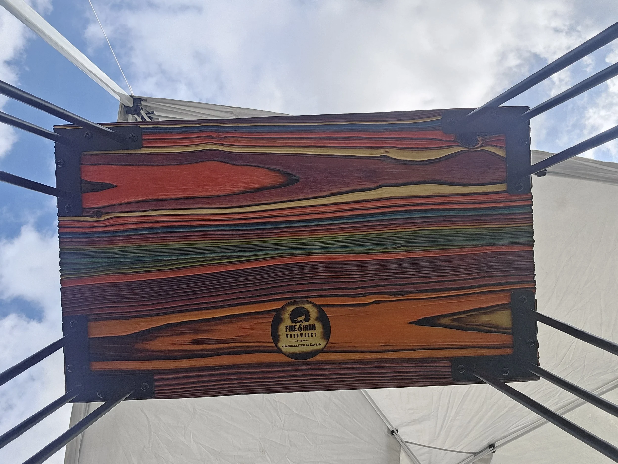 Colorful fabric hanging from a structure with black metal supports, against a cloudy sky background. There is a small round logo at the bottom with text reading "FIRE & IRON FOODVIEWS - HANDCRAFTED BY RAVEN."