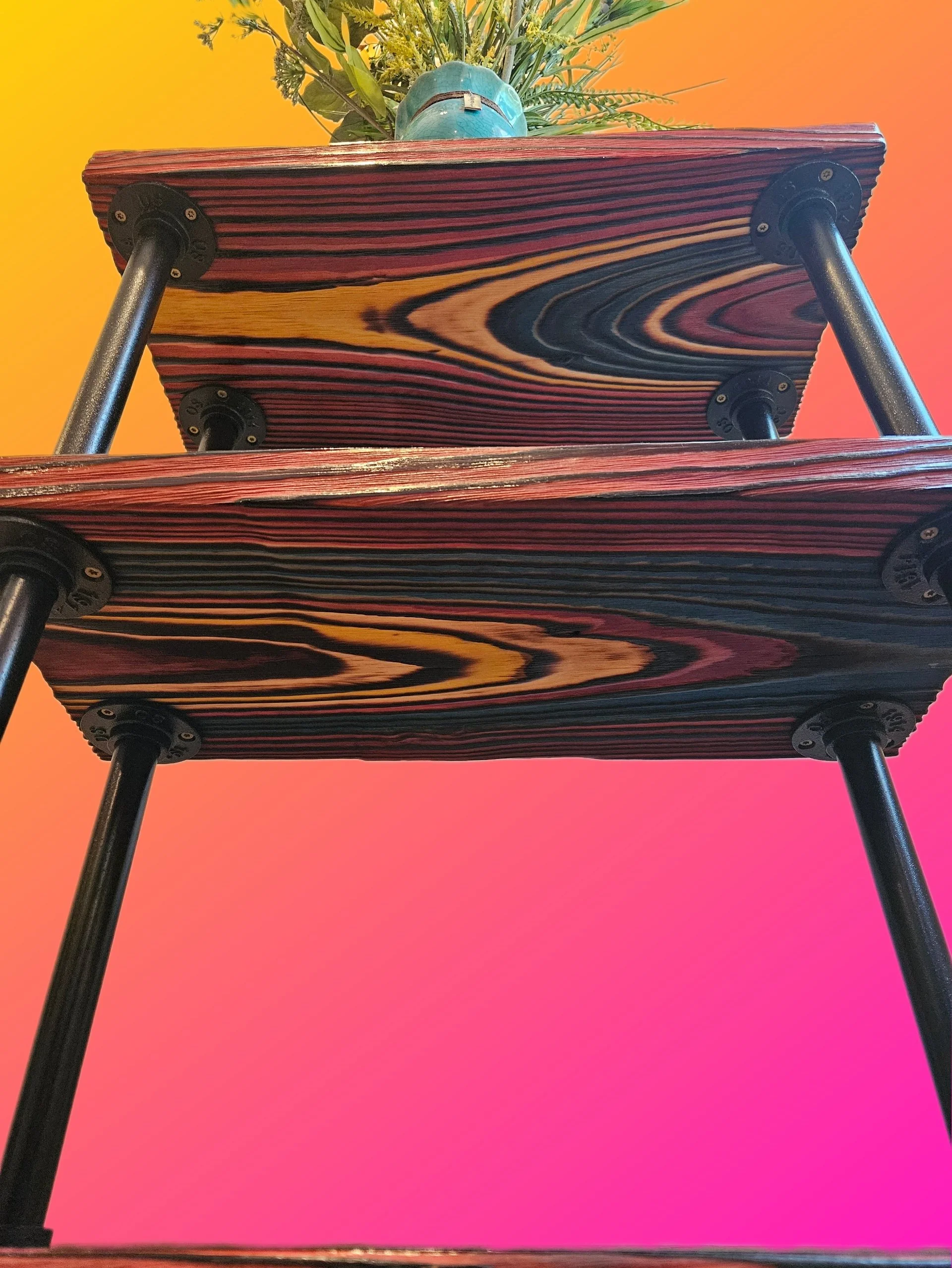 Low-angle view of a wooden table with a floral arrangement, a blue helmet, and black metal legs, against a bright orange and pink gradient background.