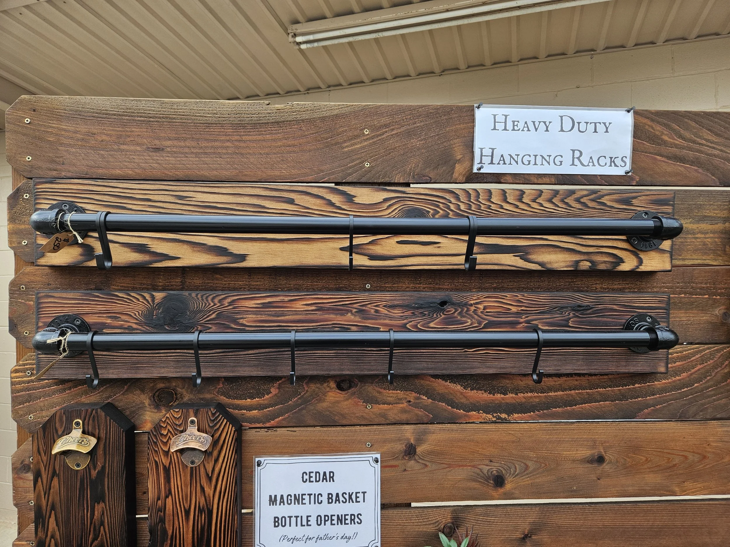 A wooden display rack labeled 'Heavy Duty Hanging Racks' with black metal bars for hanging items. Below, a sign reads 'Cedar Magnetic Basket Bottle Openers' with small decorative items hanging on the side.