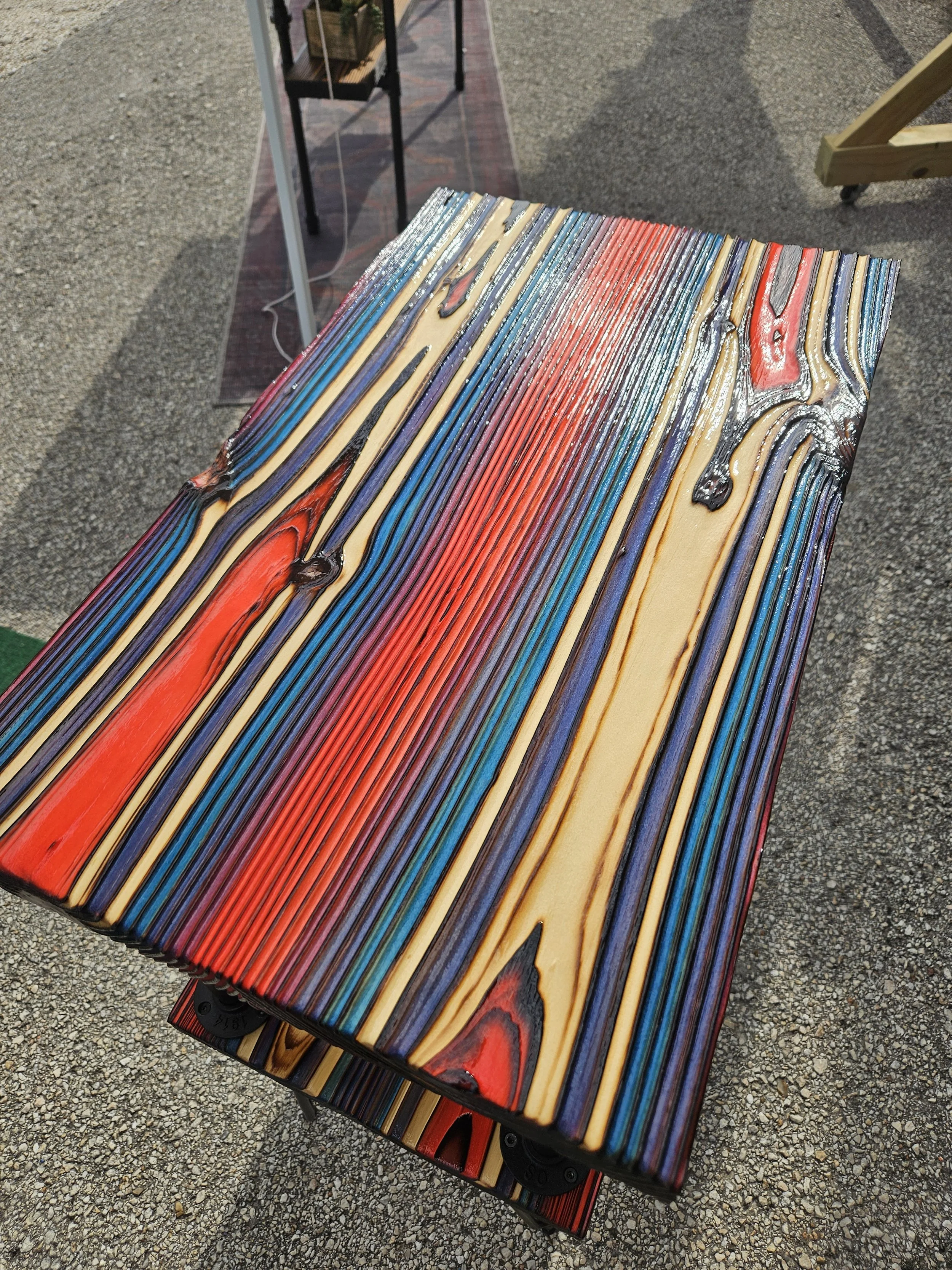 A wooden park bench with a colorful, striped pattern on the seat and backrest, situated outdoors on a gravel surface.