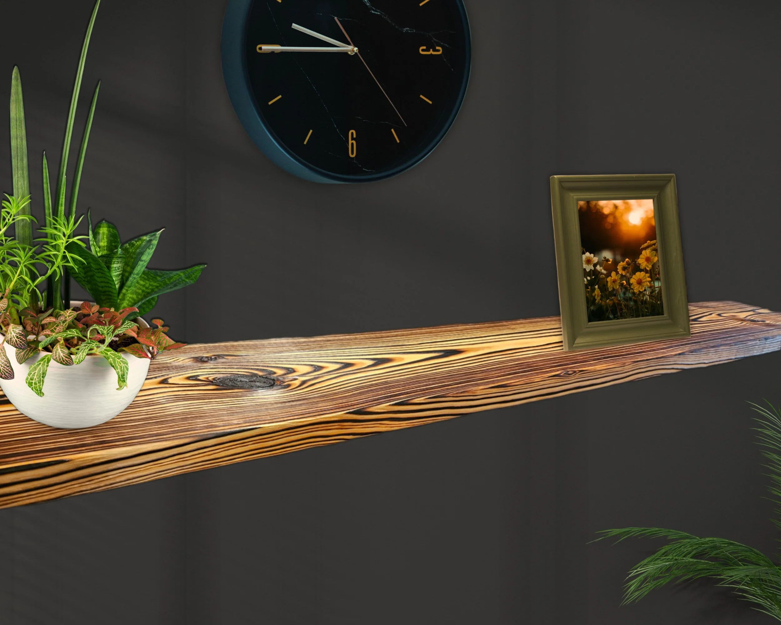 A wall clock with a black face and yellow numbers, showing approximately 12:55, hangs on a dark wall. A picture frame with a sunset and flowers reflection leans against the wall. On a wooden shelf, there are potted green plants with striped and round